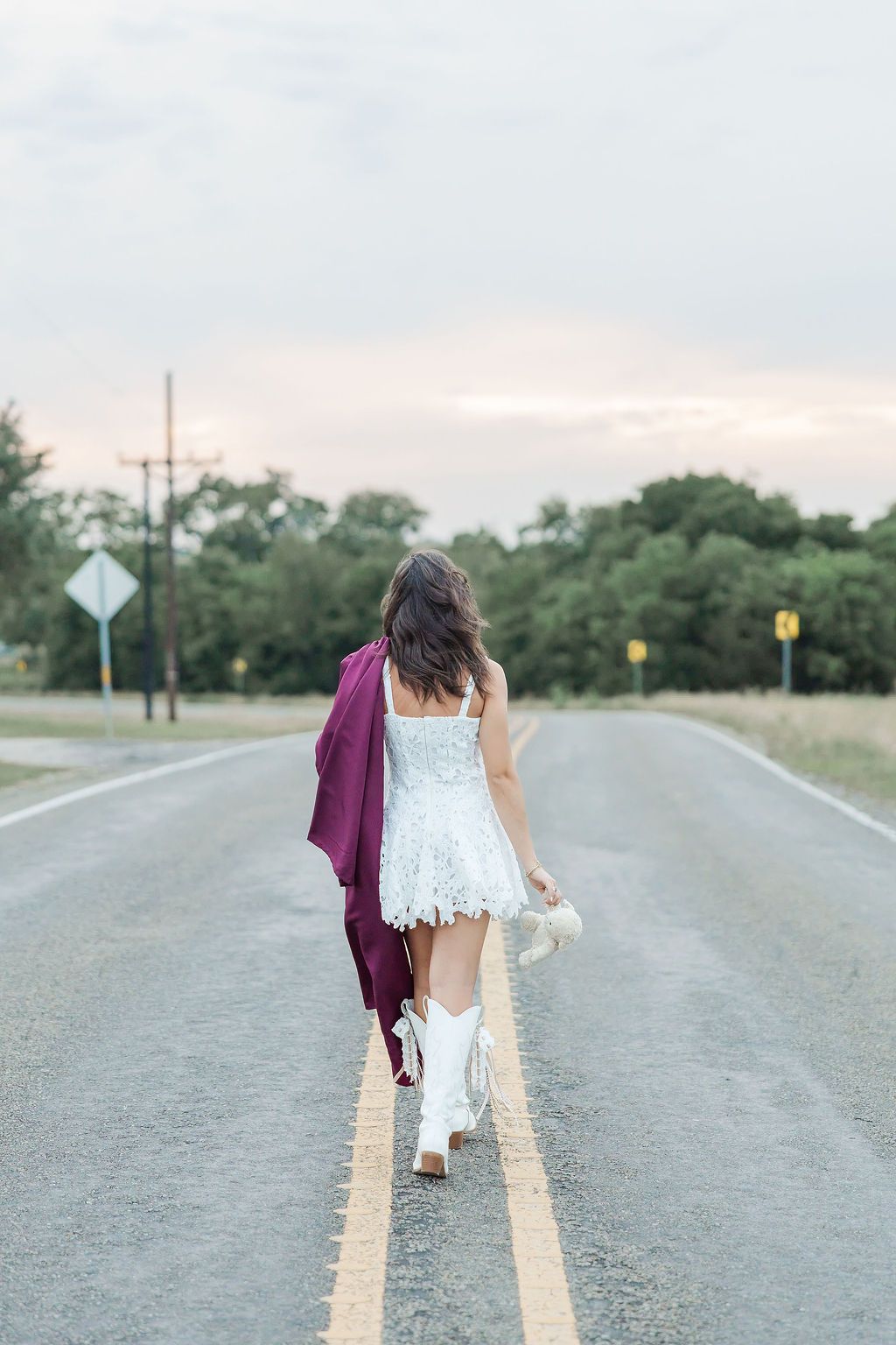 A young woman in a white lace dress and cowboy boots walks down a rural road holding a maroon graduation gown.