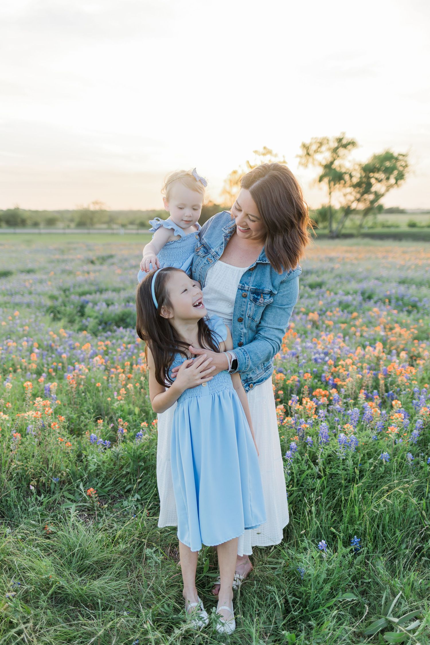 A mother holds her toddler while her daughter laughs in a bluebonnet field during a session with a Houston family photographer.