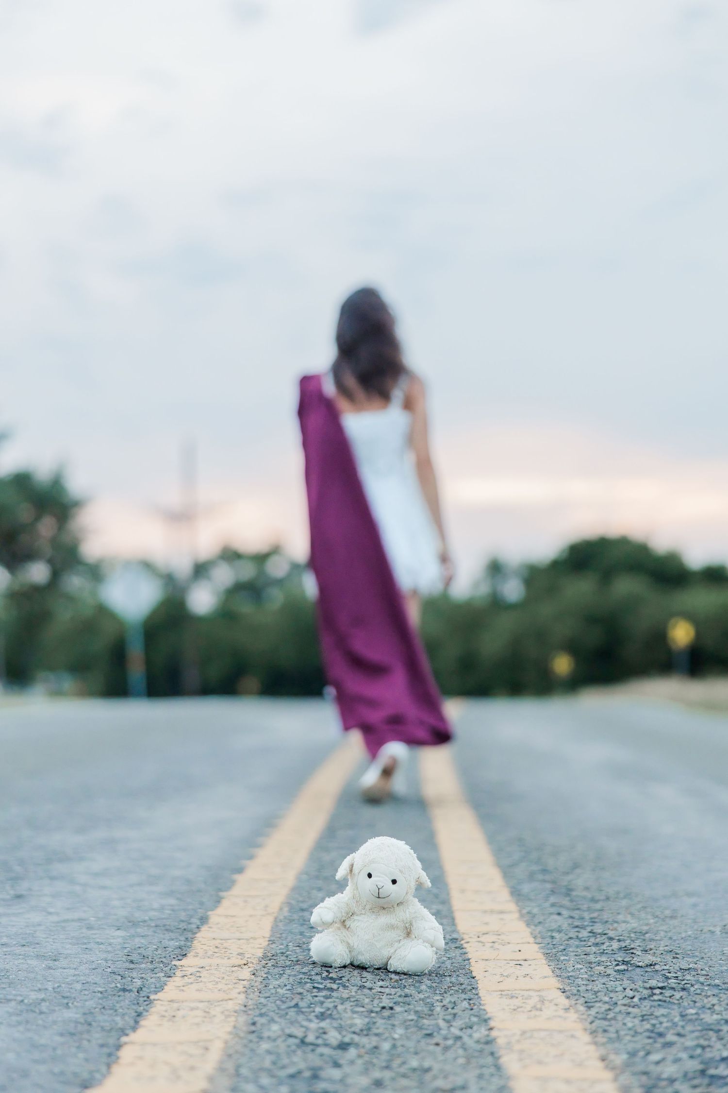 A  teddy bear sits on road markings as a senior girl with a graduation gown walks away at dusk during a Houston senior photography session.