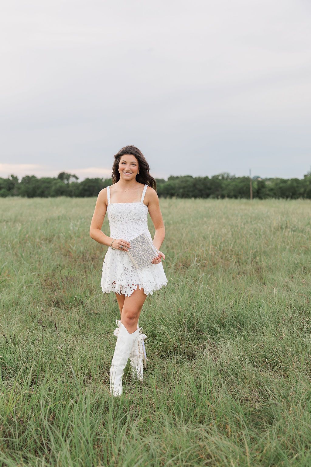 Young woman in white lace dress and cowboy boots smiling in a green field for Houston senior pictures.