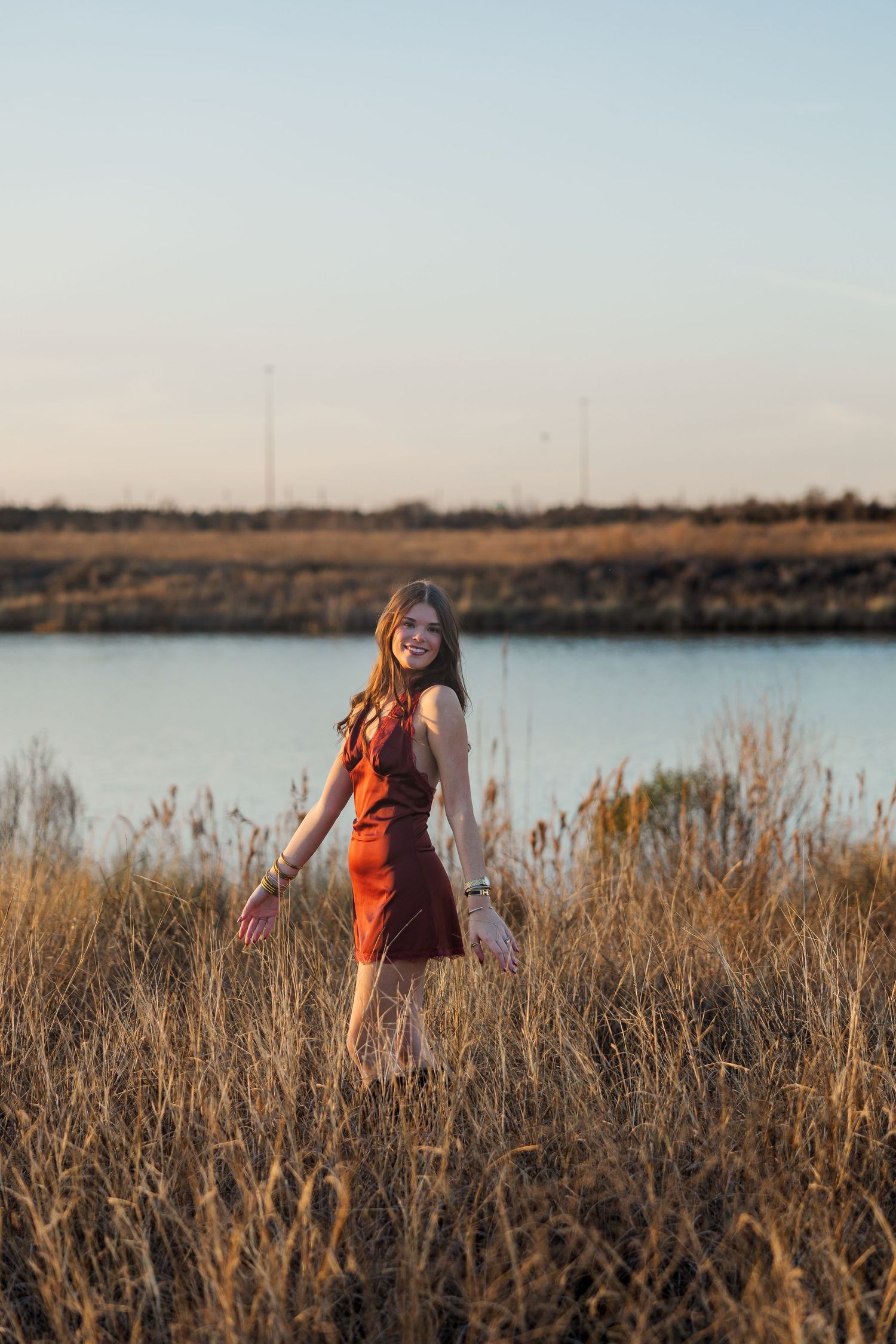 Young woman in red dress standing in golden field at golden hour during a session with a Houston senior photographer.