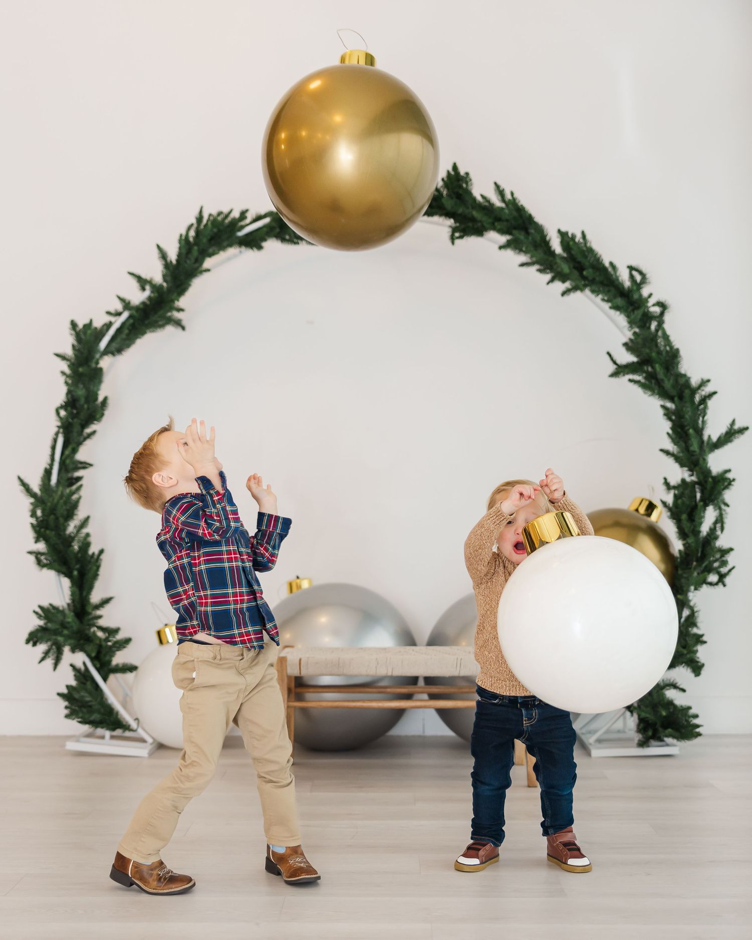 Two children play with oversized gold  ornaments in front of a festive green wreath arch holiday backdrop for a Christmas photoshoot houston