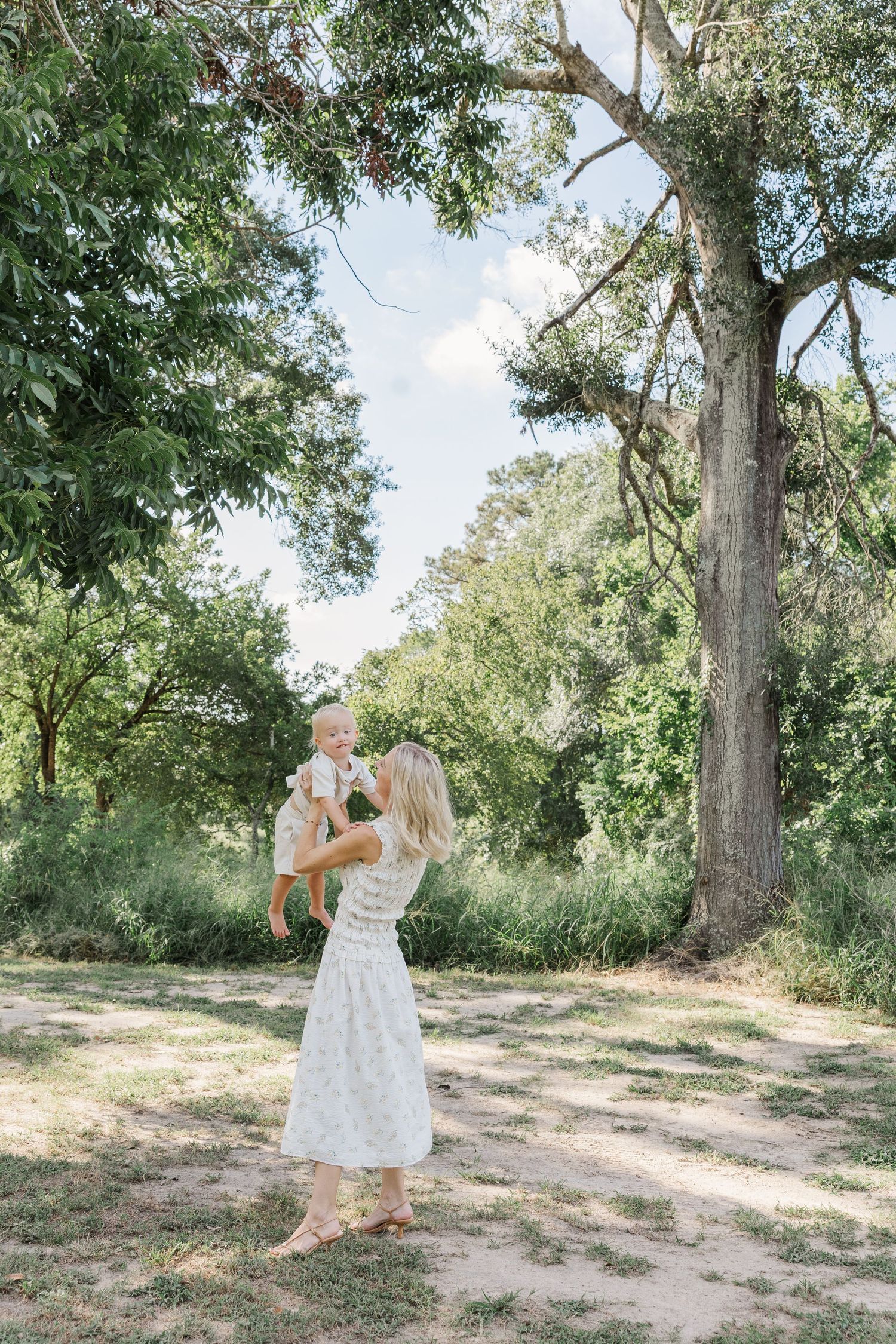 A woman in a white dress lifts a baby outdoors surrounded by lush green trees  during a session with a Houston newborn photographer.