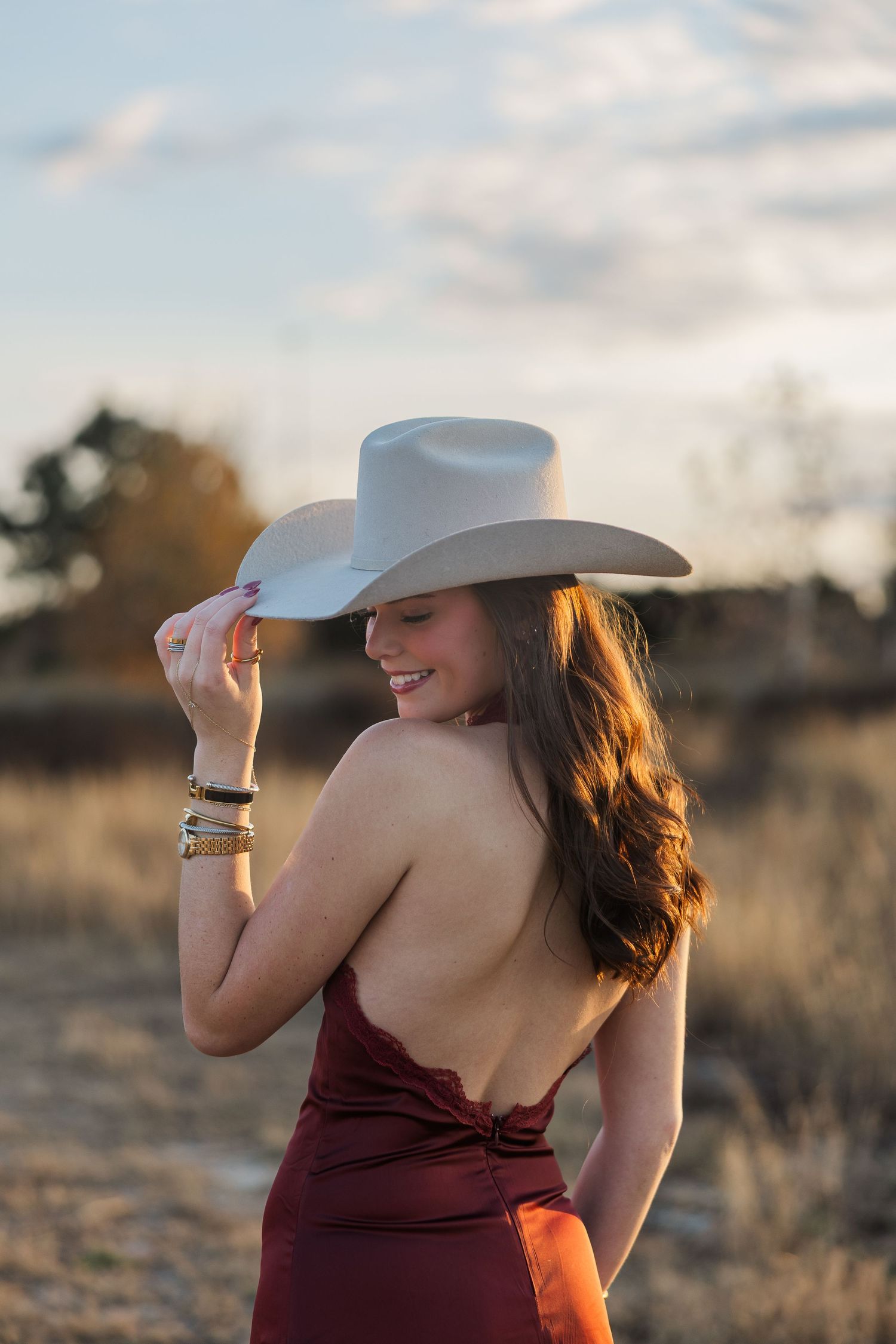 Young woman in backless red dress and white cowboy hat smiling outdoors during senior pictures in Houston at golden hour.