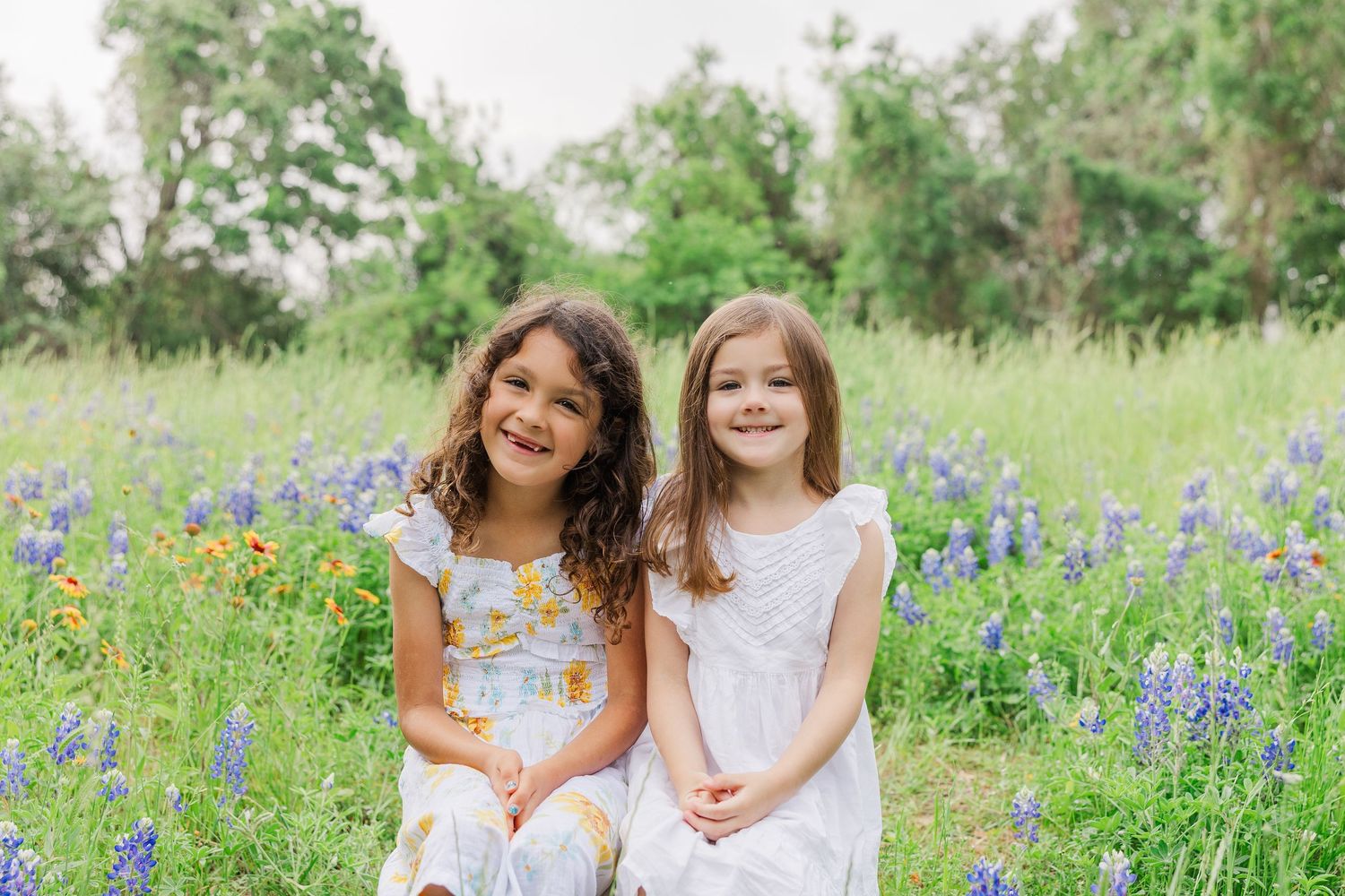 Two young girls in white dresses sit together in a field of blooming bluebonnets.