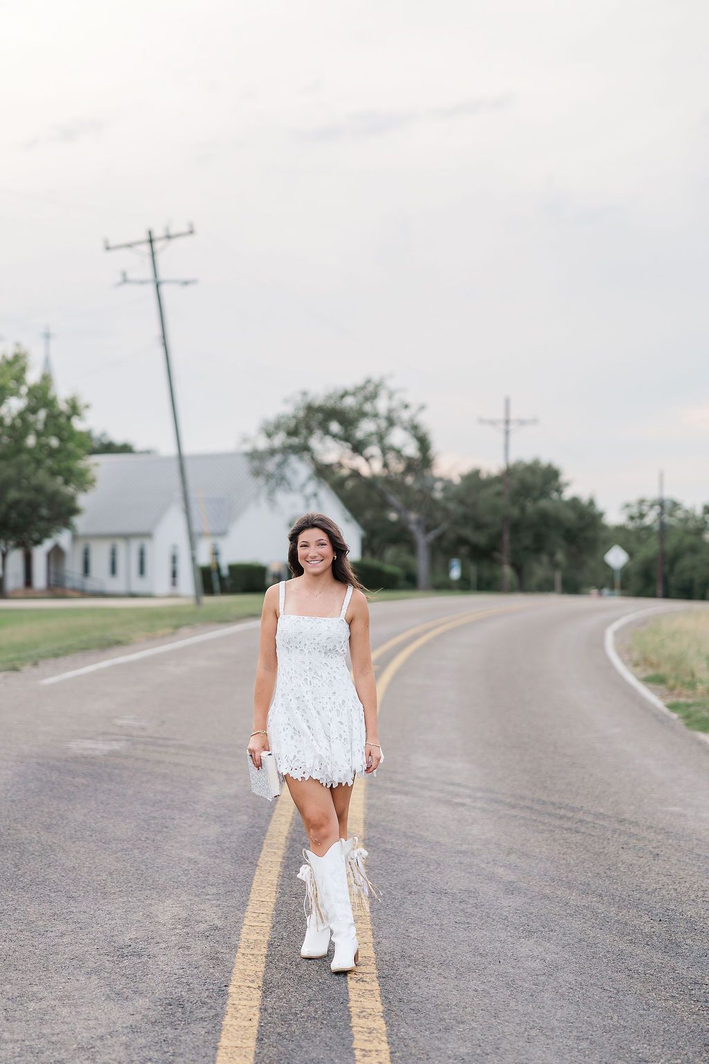 A photo taken by a Houston senior photographer of a woman in a lace mini dress and cowboy boots smiling while walking on a rural Texas road.