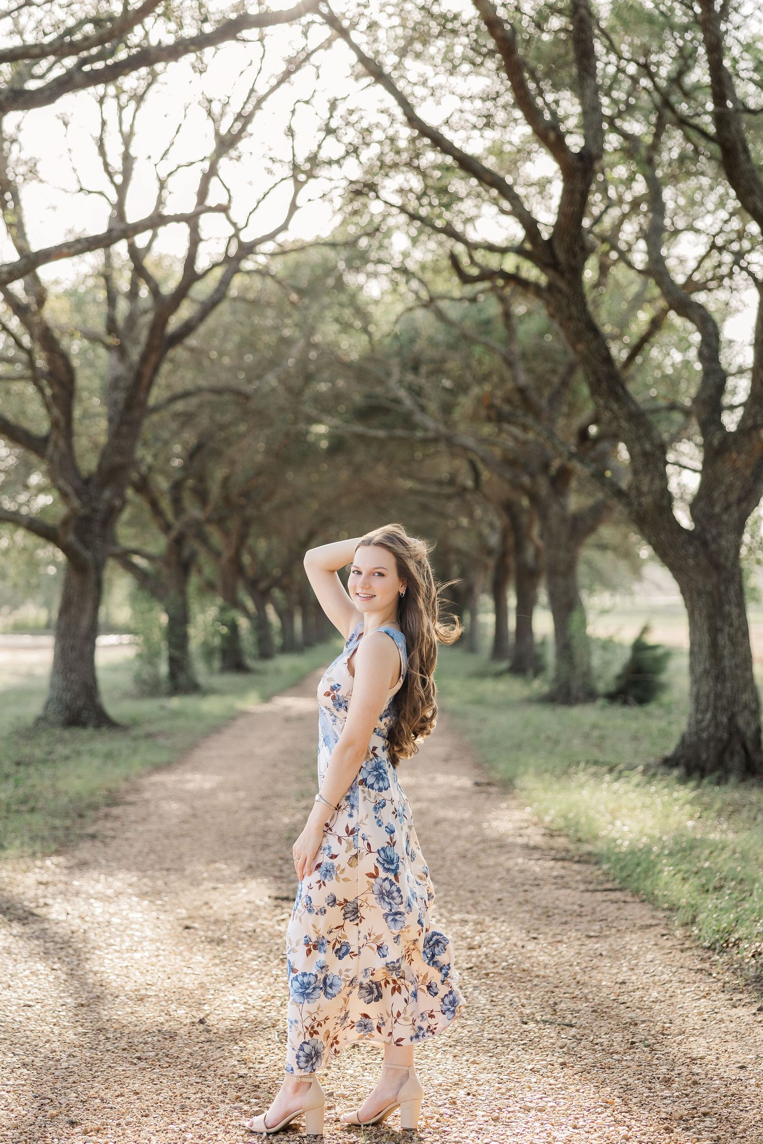 Young woman in floral dress poses on a sunlit tree-lined dirt path, smiling during a Houston senior photography session.