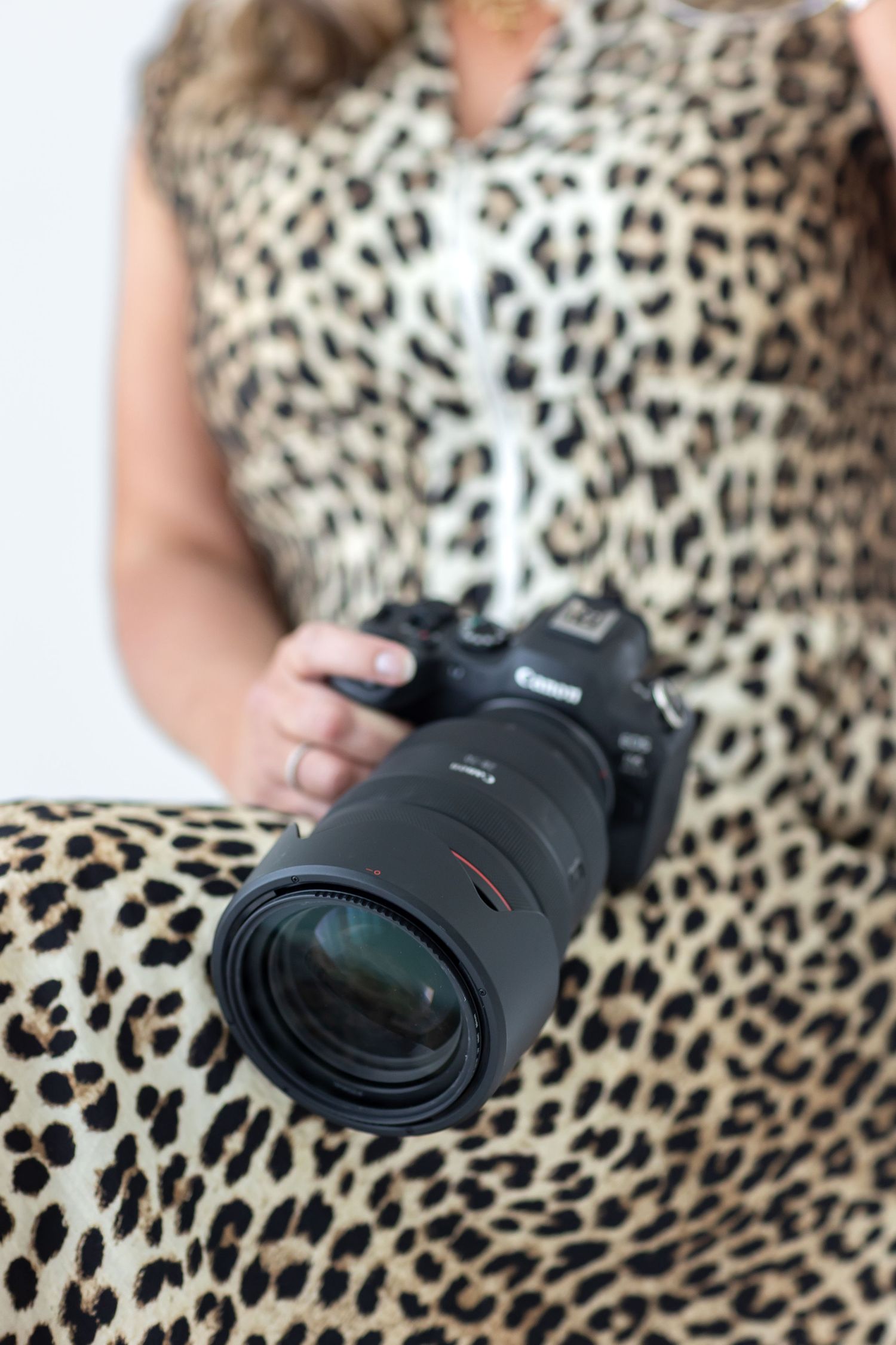 A Houston senior photographer in a leopard print dress holding a Canon DSLR camera with a large telephoto lens in focus foreground.