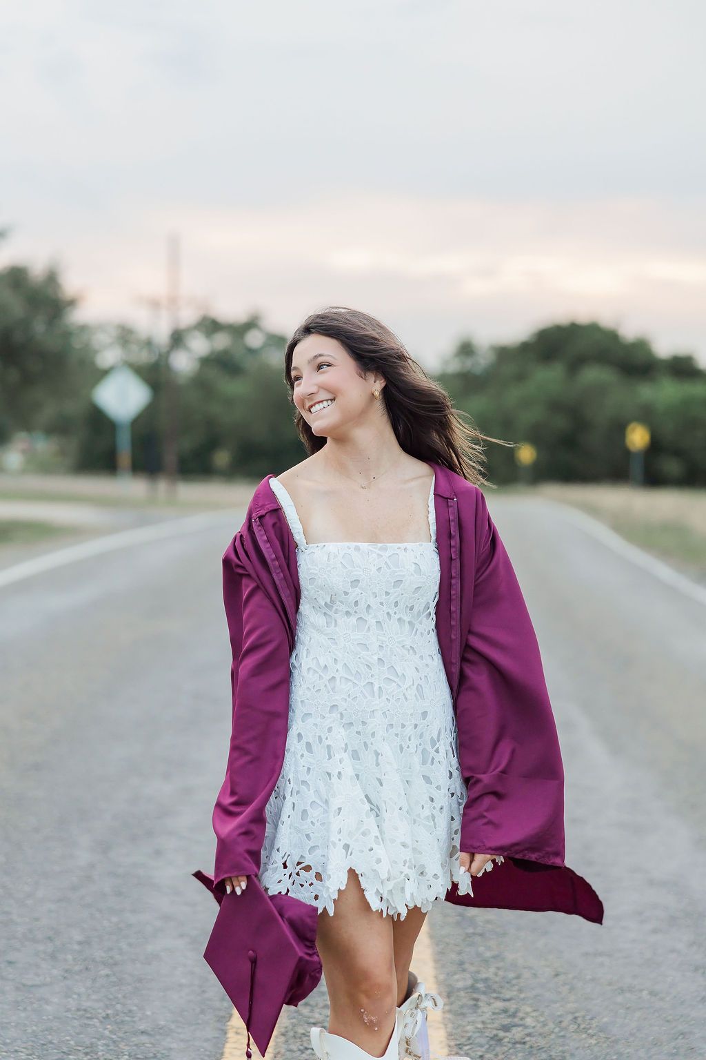 Smiling young woman in white lace dress and maroon graduation gown standing on open road at dusk during a Houston Texas senior photoshoot.