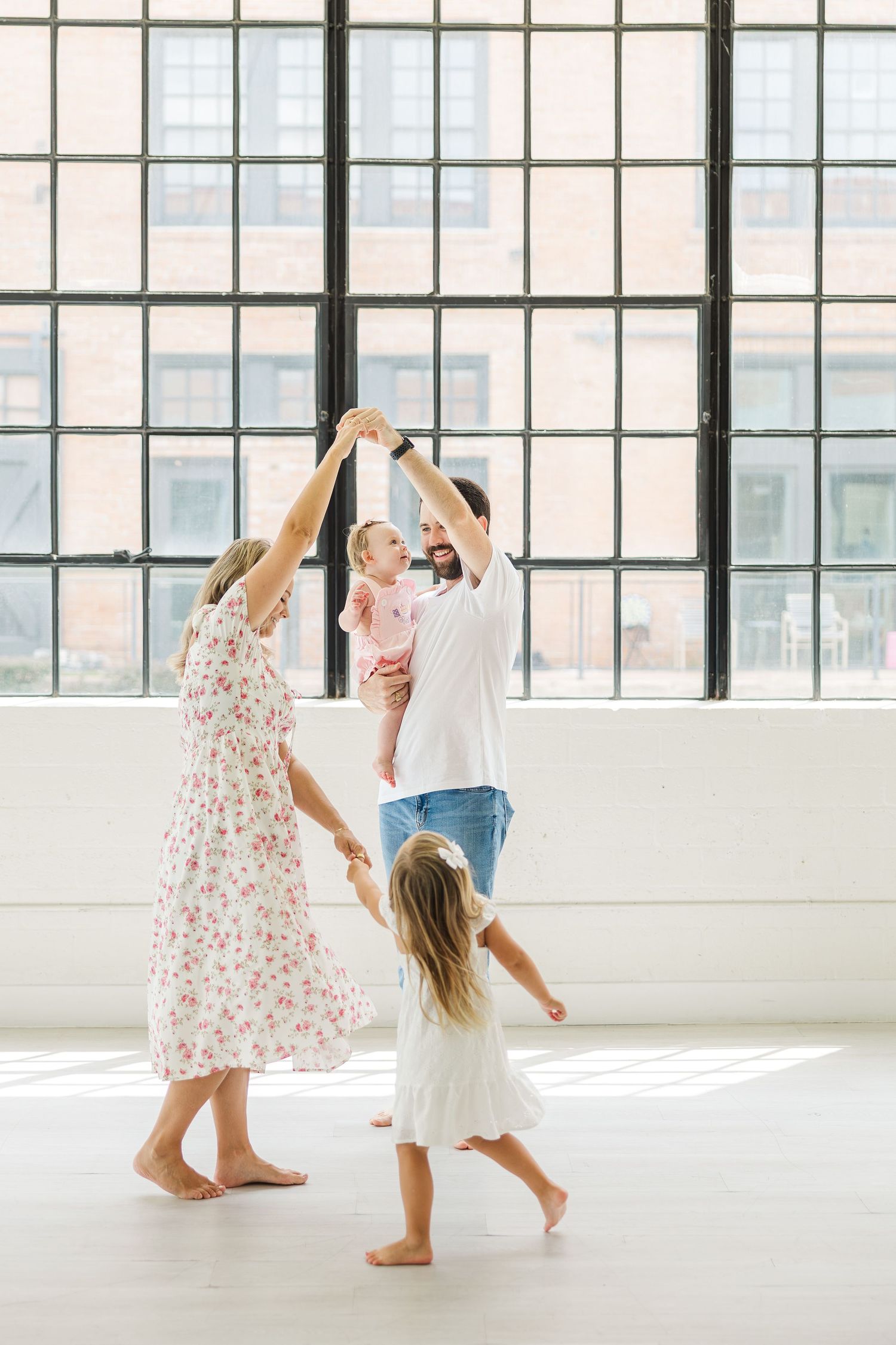 A joyful family dancing together in a sunlit studio with large windows, parents lifting children playfully for Houston family pictures.