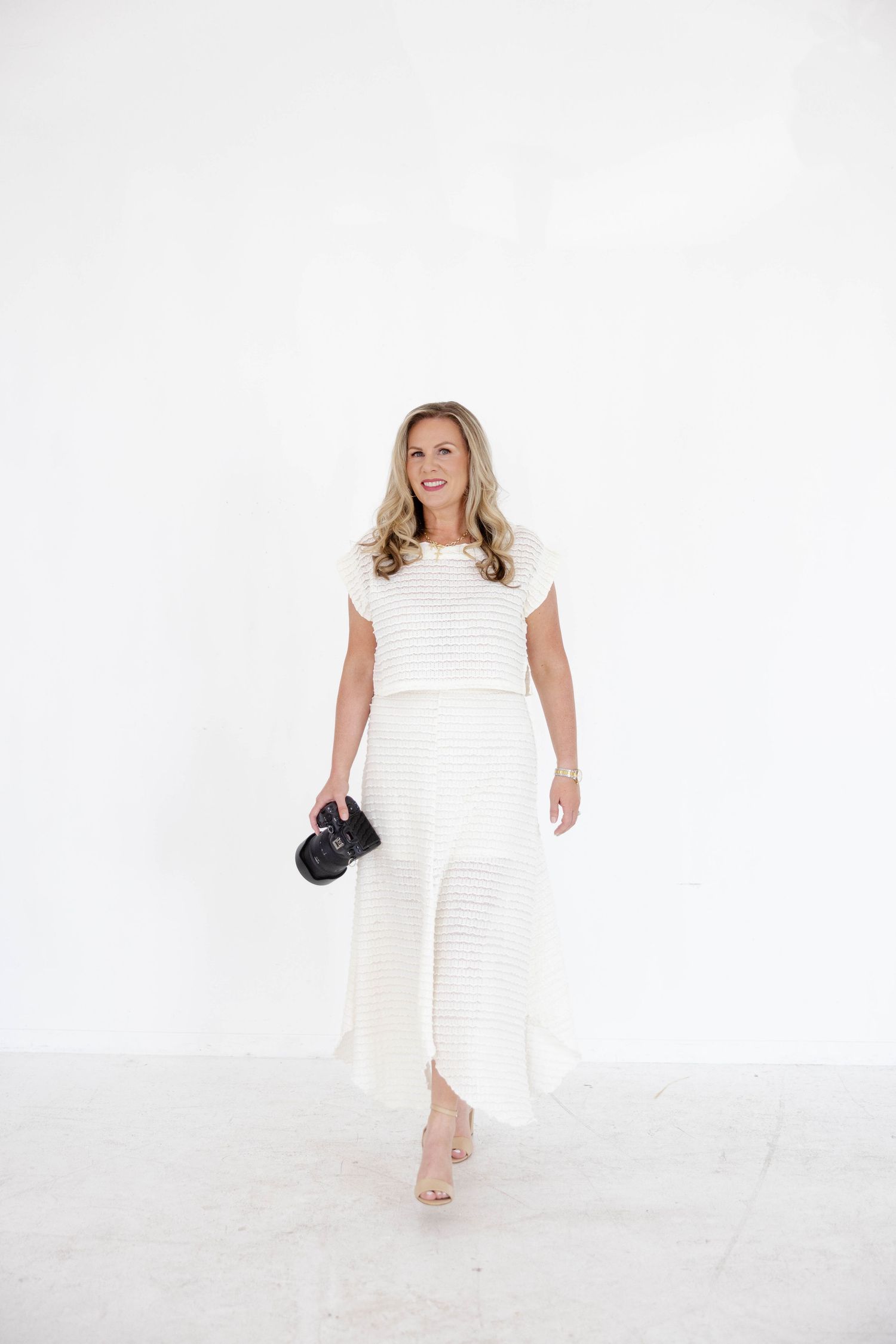 Woman in white striped maxi dress holding black clutch, walking barefoot against white background.