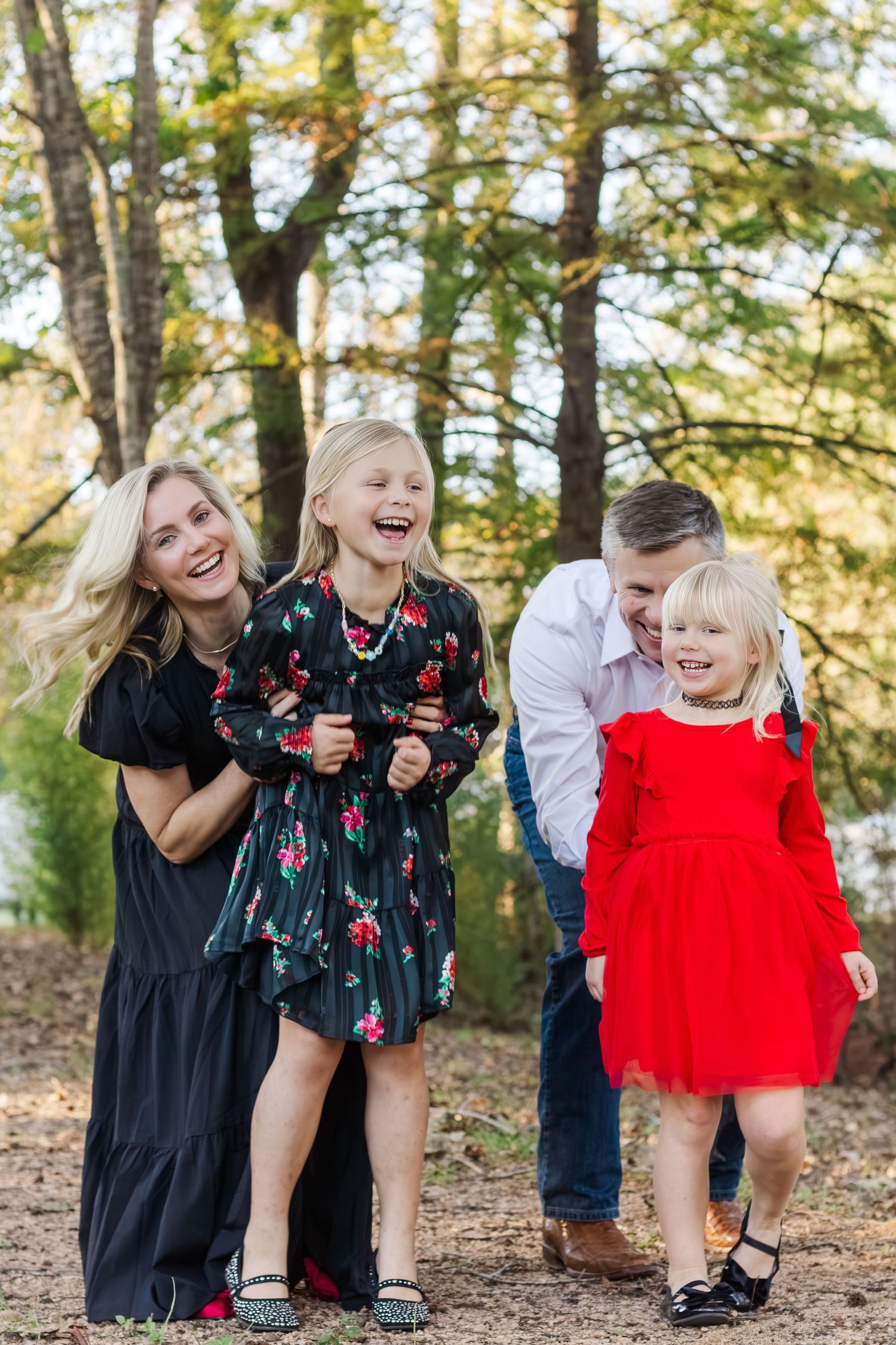 Happy family of four laughing outdoors in fall, two girls in floral and red dresses with smiling parents for Houston Christmas photos.