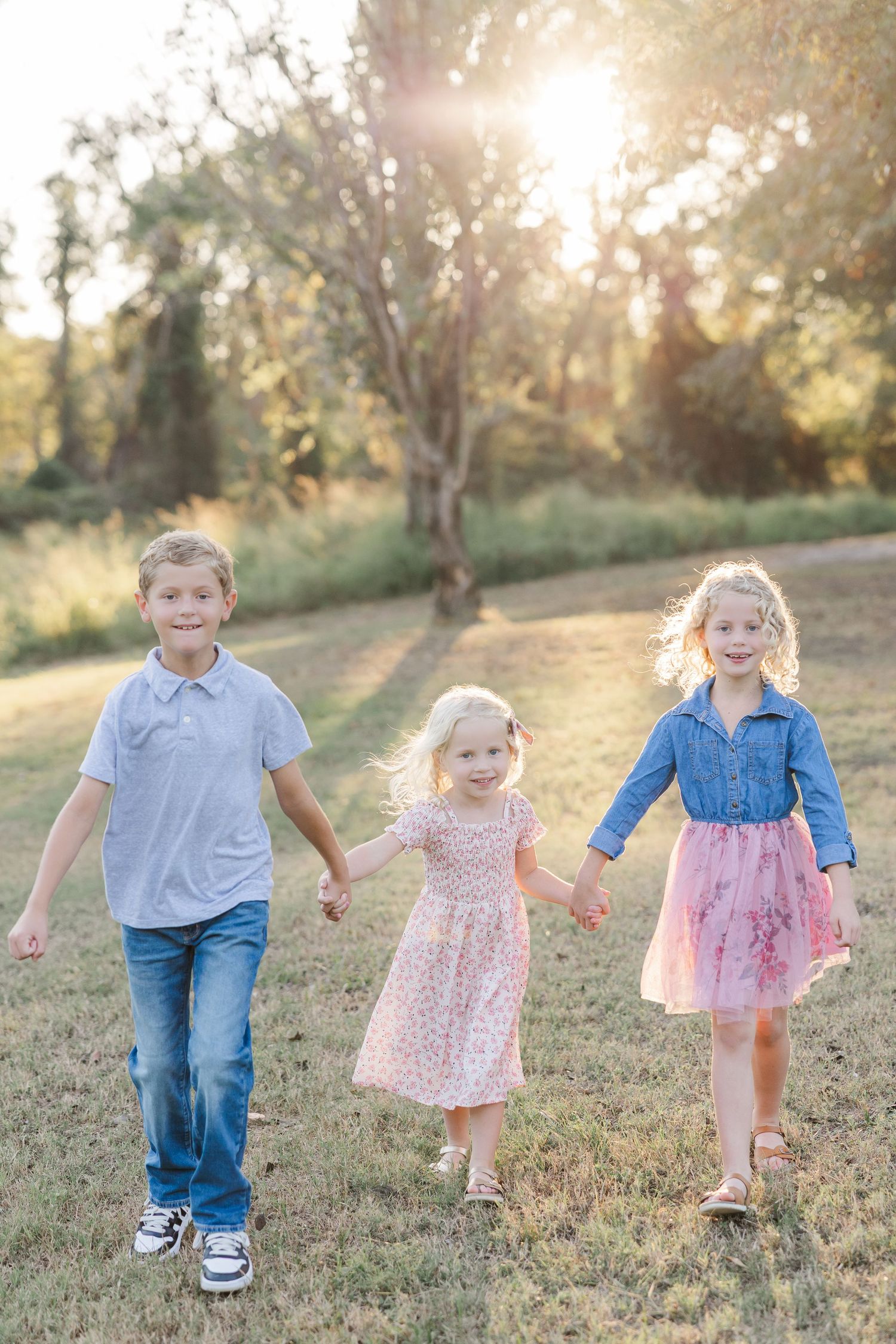 Three children holding hands and walking through a sunny park during a photoshoot with a Houston newborn photographer.