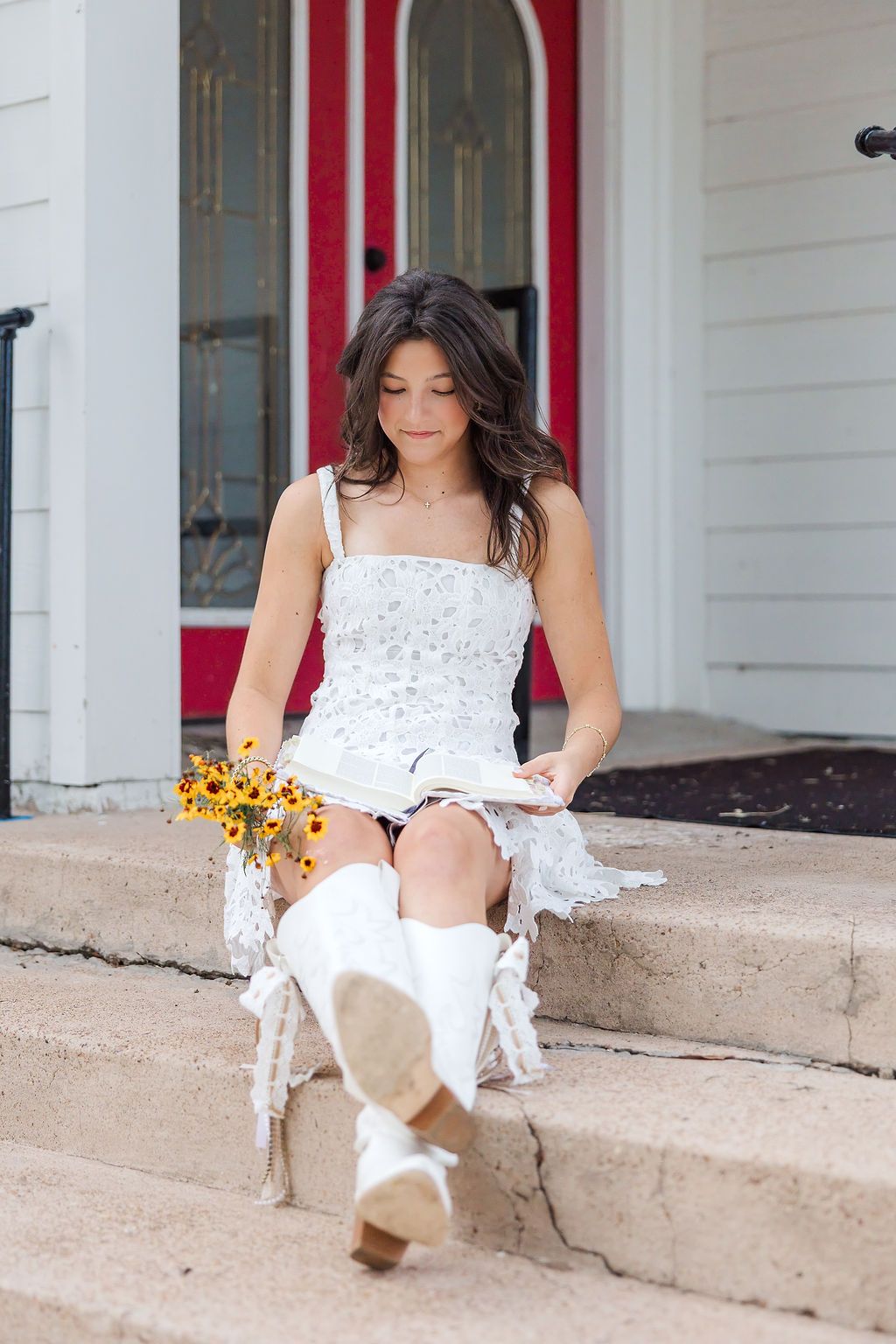 Young woman in white floral dress sits on steps reading a Bible during a session with a Houston senior photographer.