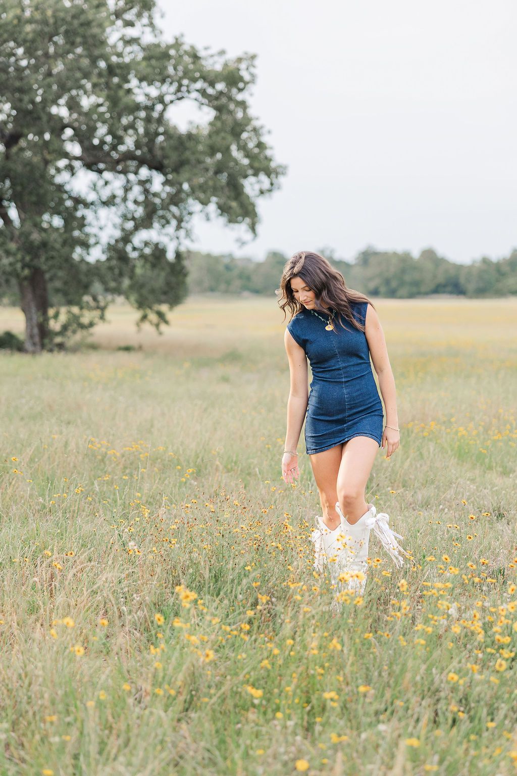 A young woman in a blue mini dress and white boots walks through a wildflower meadow during senior pictures in Houston TX.