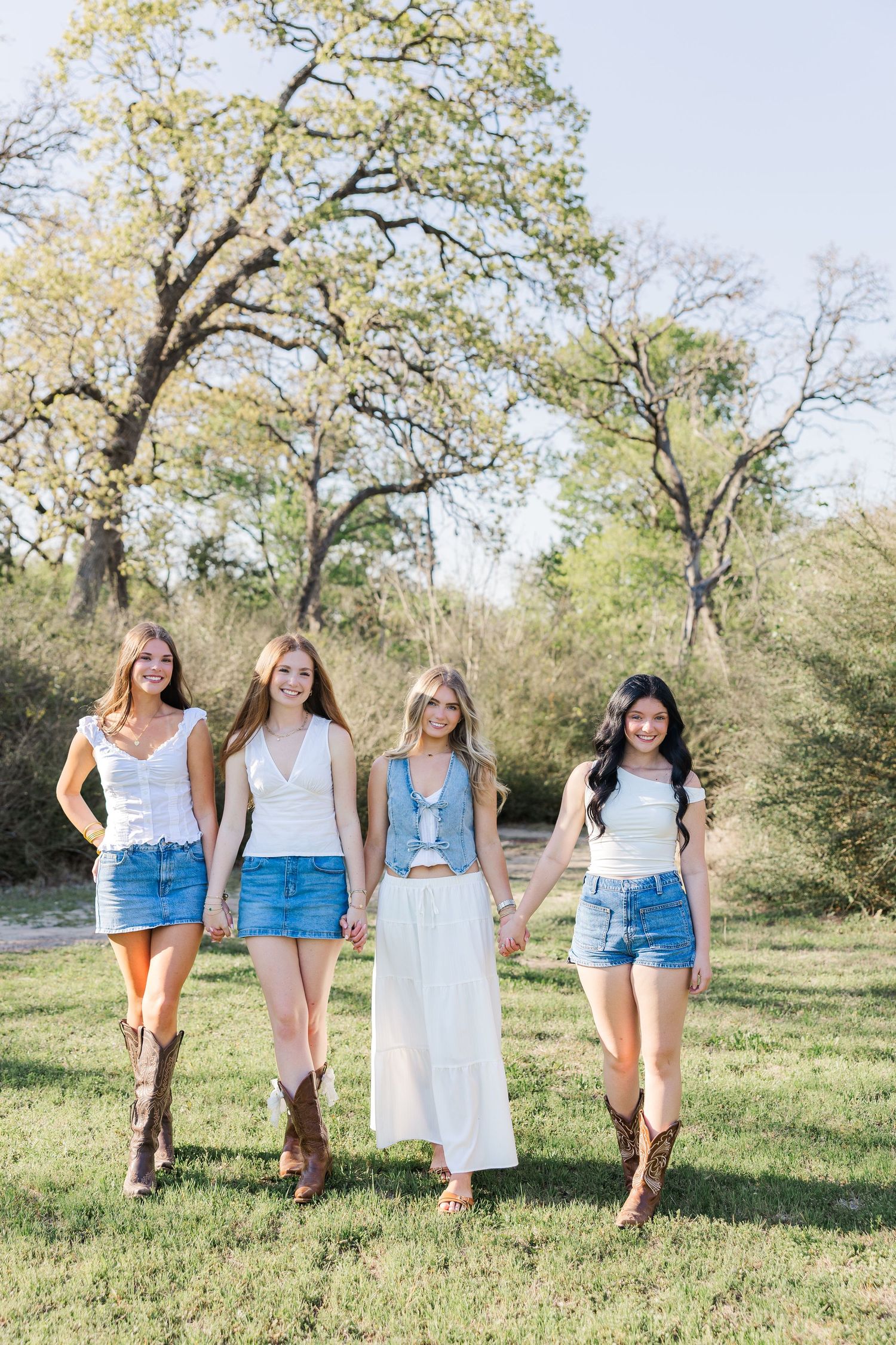 Four young women in white tops, denim shorts, and cowboy boots hold hands walking outdoors during a Houston senior photography session.