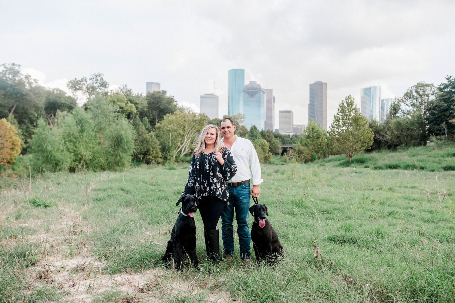 A couple stands with their two black dogs against a city skyline backdrop in a grassy field.