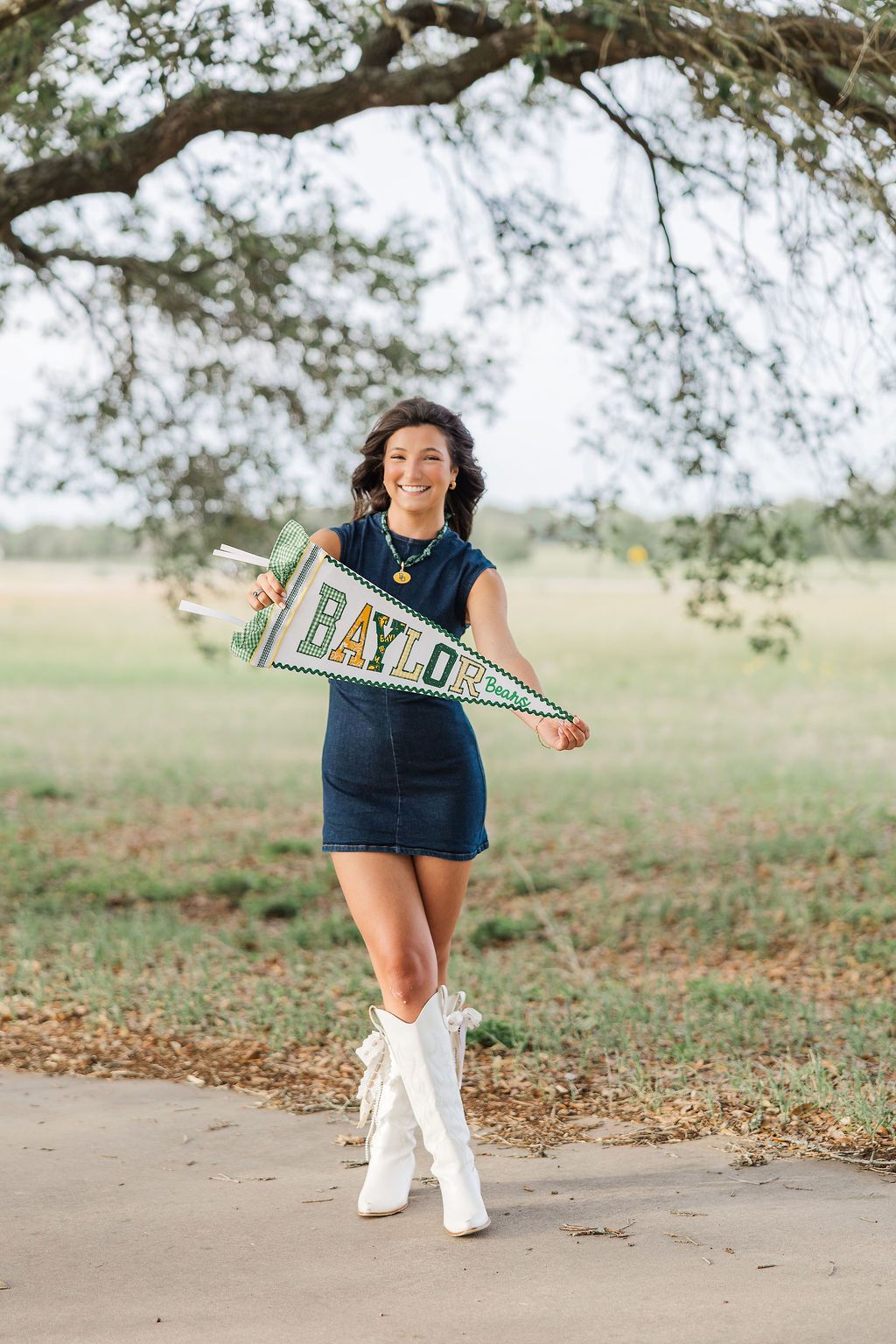 Smiling young woman in denim dress and white boots holding a Baylor University pennant flag outdoors.