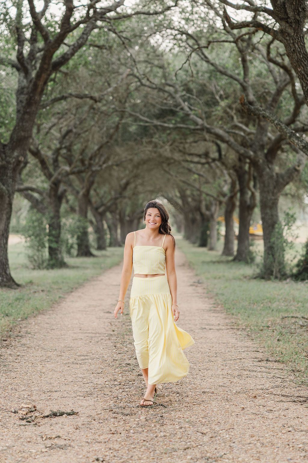 Young woman in yellow crop top and maxi skirt walking down a tree-lined dirt path, smiling for senior photos in Houston TX.