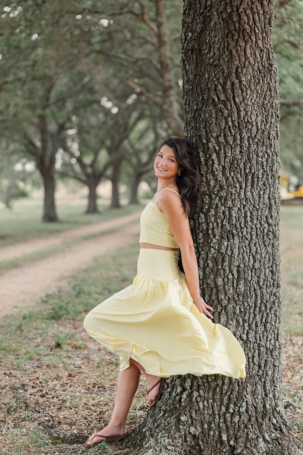 Young woman in yellow crop top and flowing skirt leaning against a large tree in a lush green park during Houston senior photos.