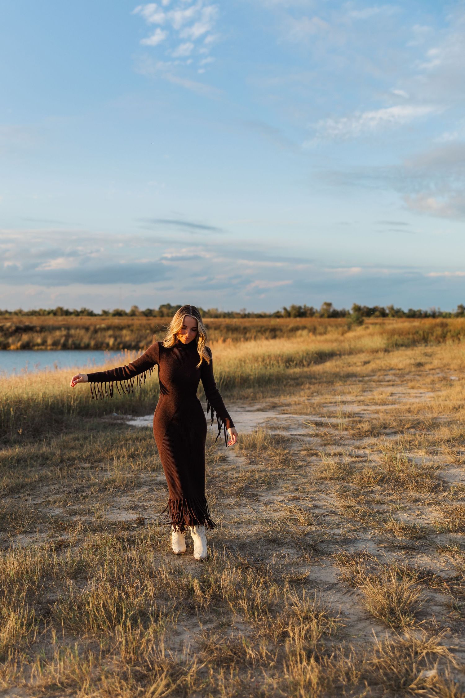 Woman in brown fringe dress and white boots poses in golden field near water during a Houston senior photography shoot.