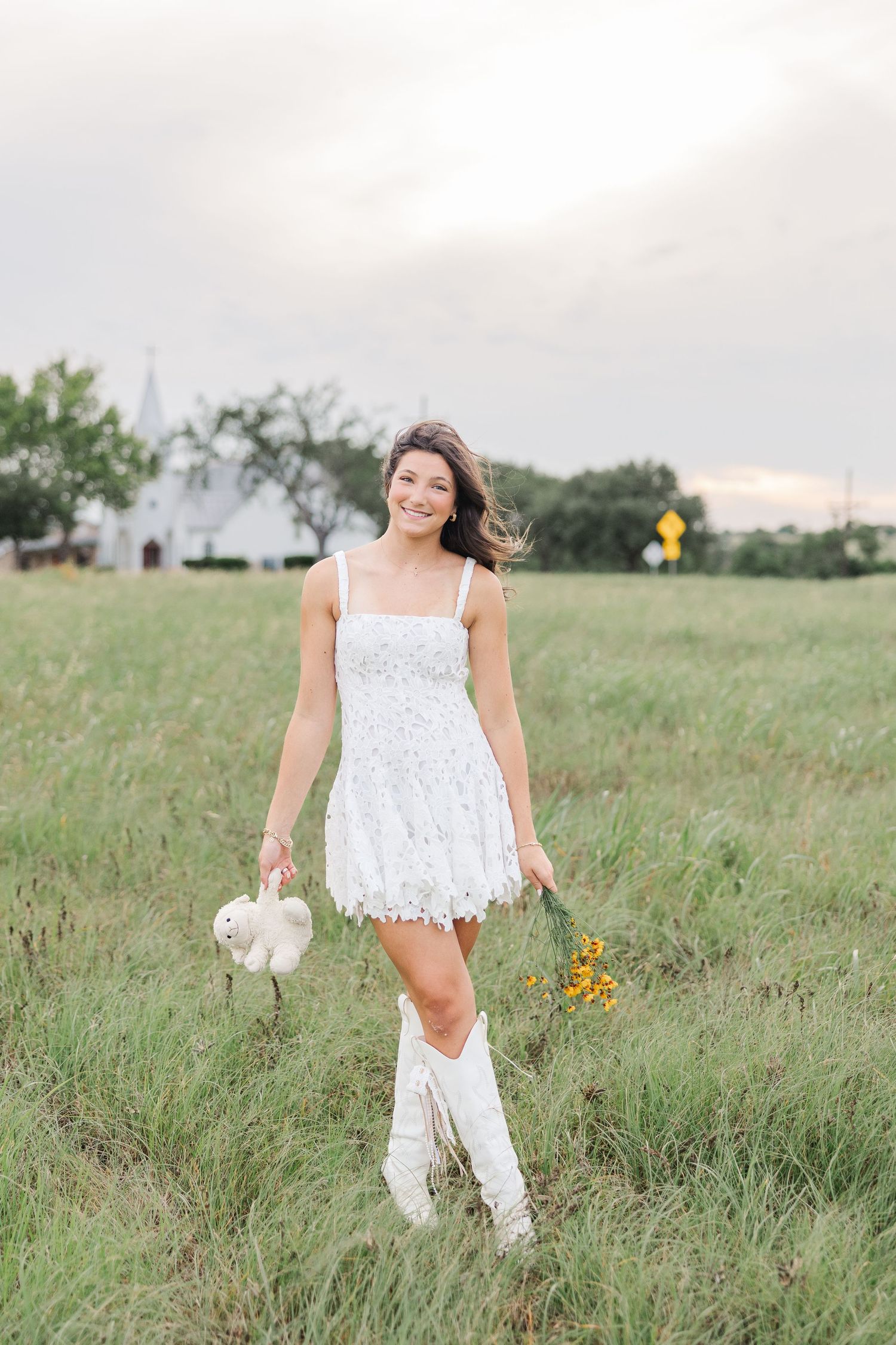 Young woman in a lace dress and cowboy boots walks through a field holding wildflowers during senior pictures in Houston.