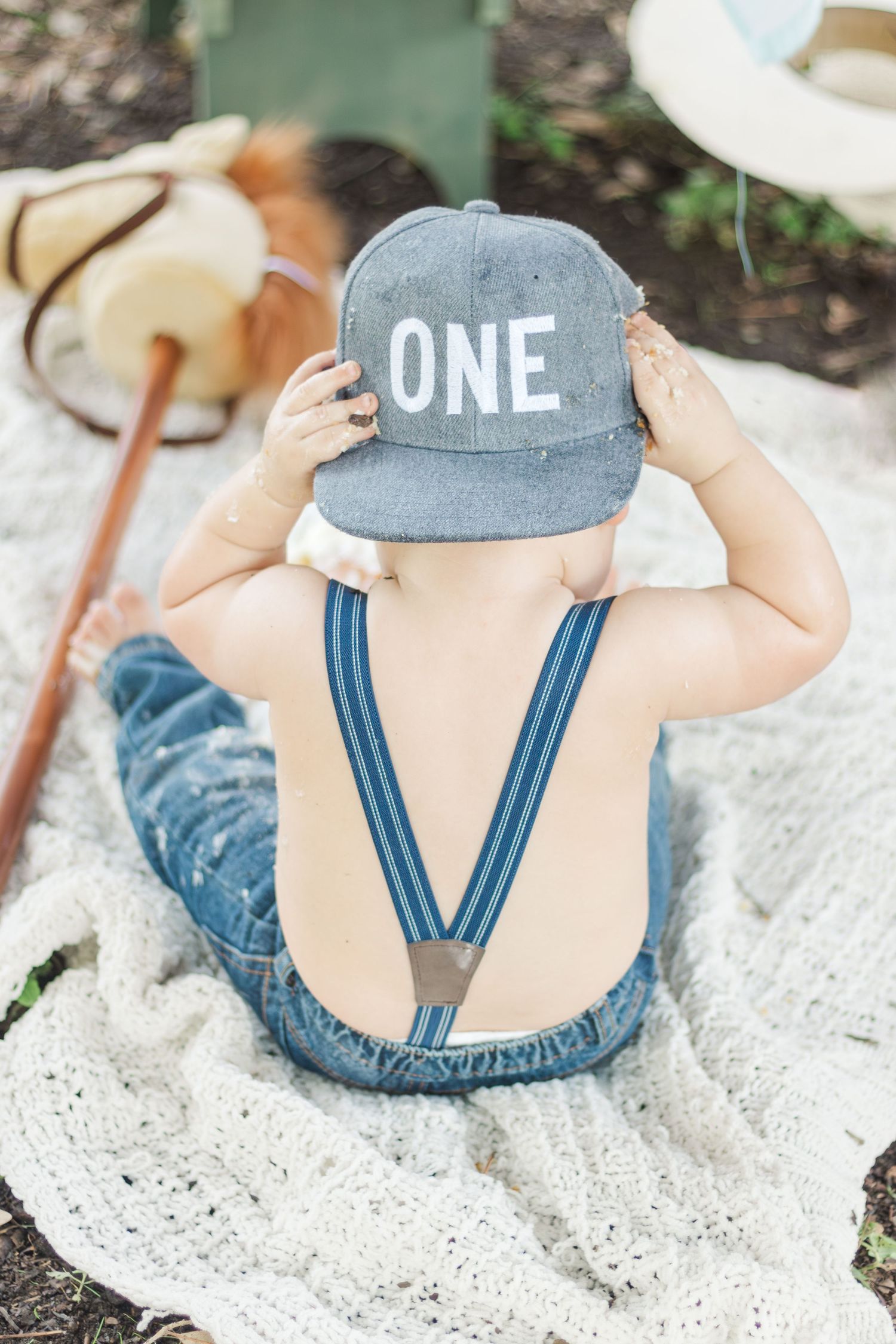 Back view of a small child wearing a gray cap with 'ONE' text and blue overalls sitting on a blanket.