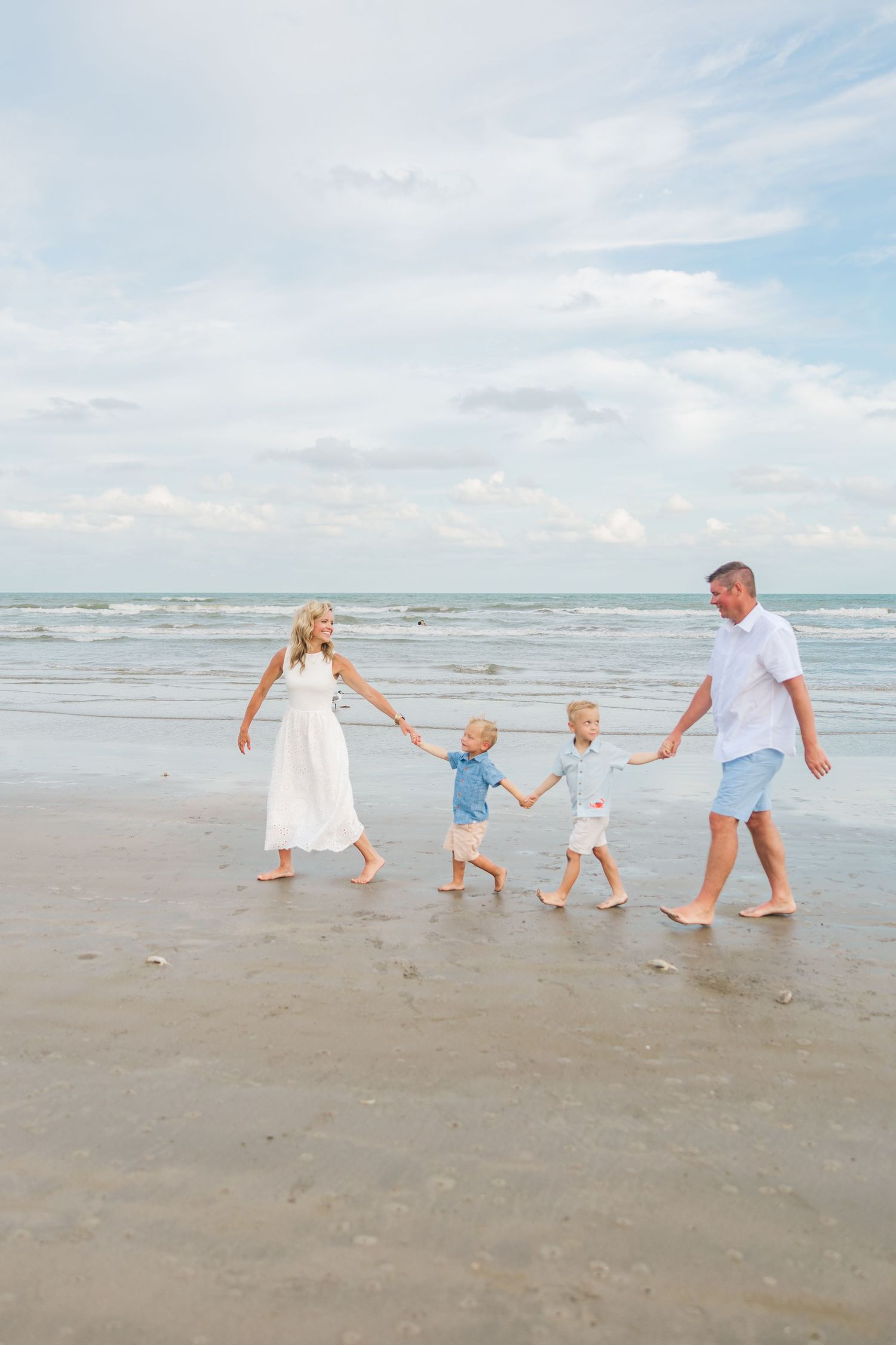 Family of four walking hand-in-hand along a sandy beach with ocean waves and cloudy sky backdrop during family photography Houston TX.