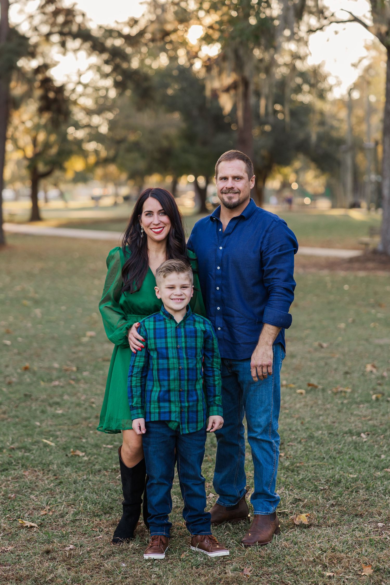 Family of three posing outdoors in a park, wearing coordinating green and blue outfits for houston christmas pictures.