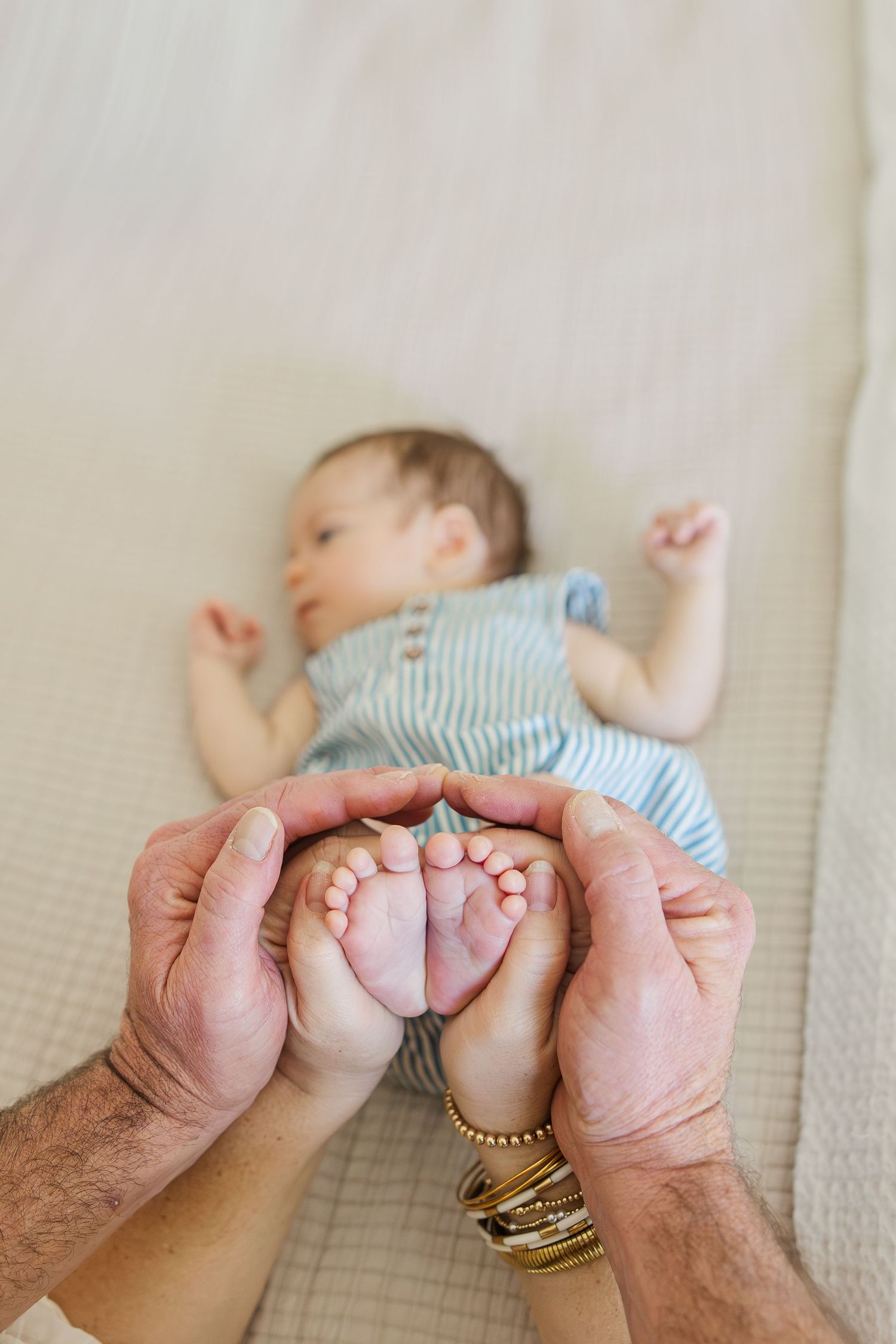 Adult hands gently cradle a newborn baby's tiny feet while the infant rests on a cream-colored blanket in striped outfit.