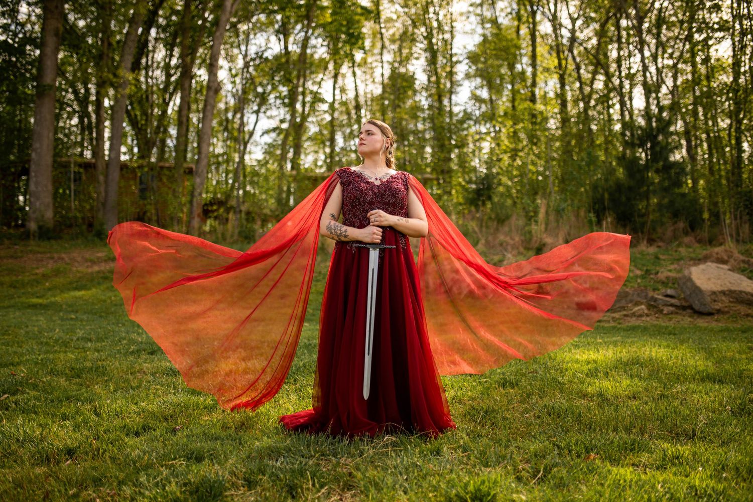 Woman in red gown with flowing orange cape holds sword outdoors among trees in fantasy-inspired photoshoot.