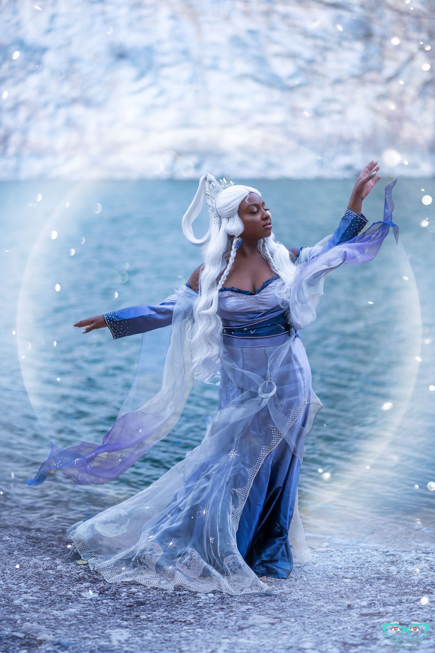 A fantasy-inspired cosplay dancer in flowing blue and white dress performs gracefully by a waterfront in ethereal lighting.