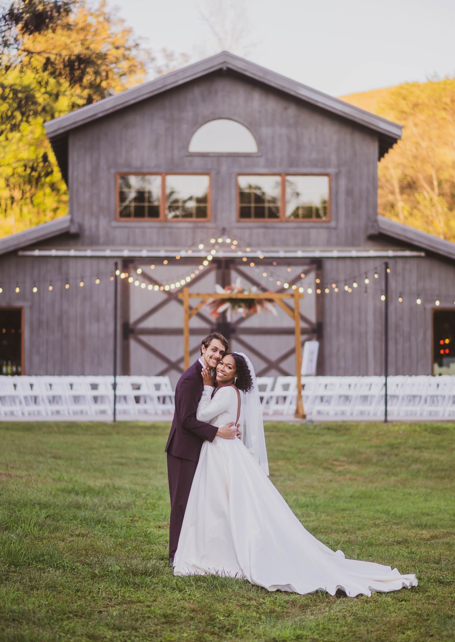 A wedding couple embraces in front of a rustic barn venue decorated with string lights at sunset.