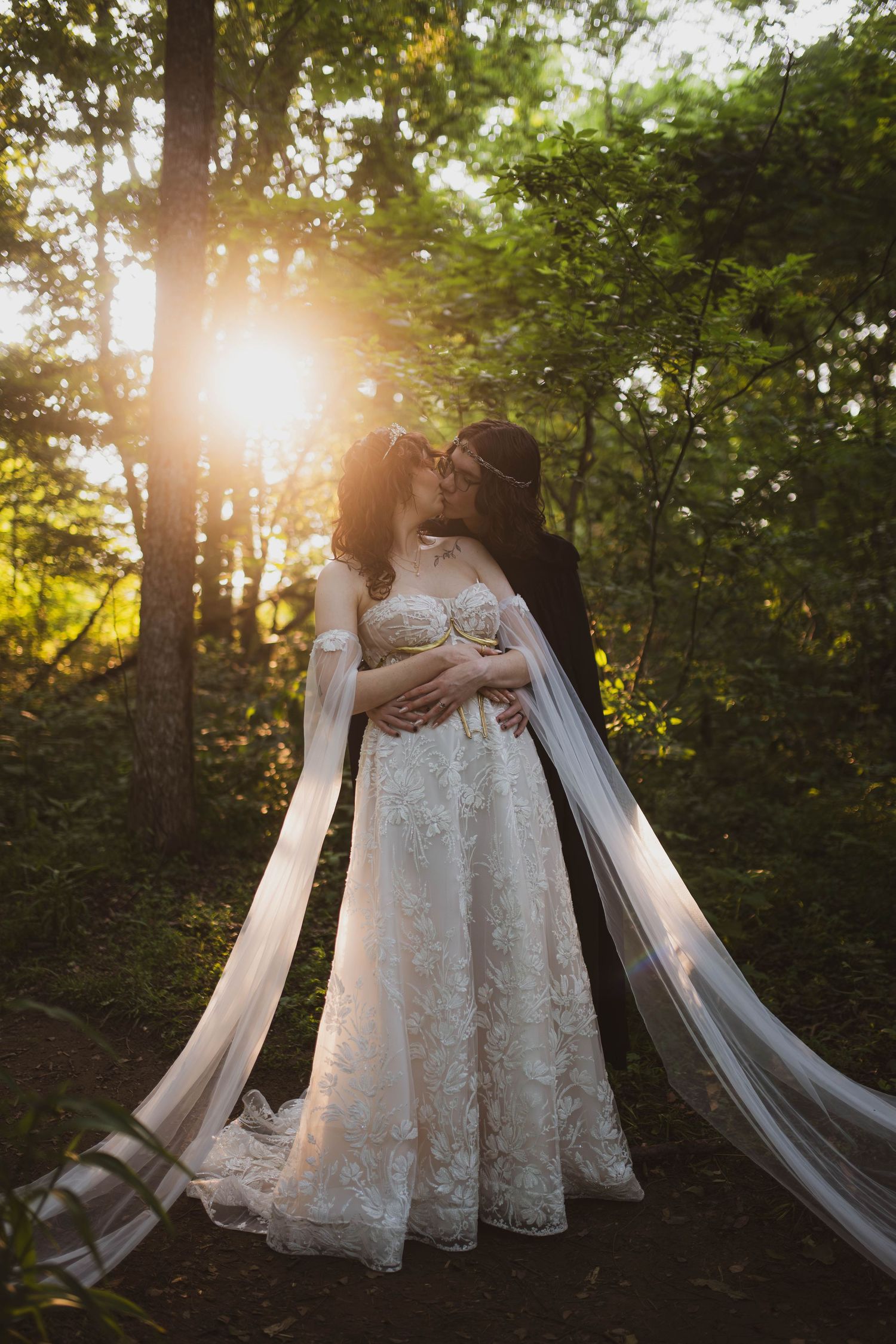A romantic golden hour wedding photo showing a couple in a white lace dress and cape embracing in a sunlit forest setting.
