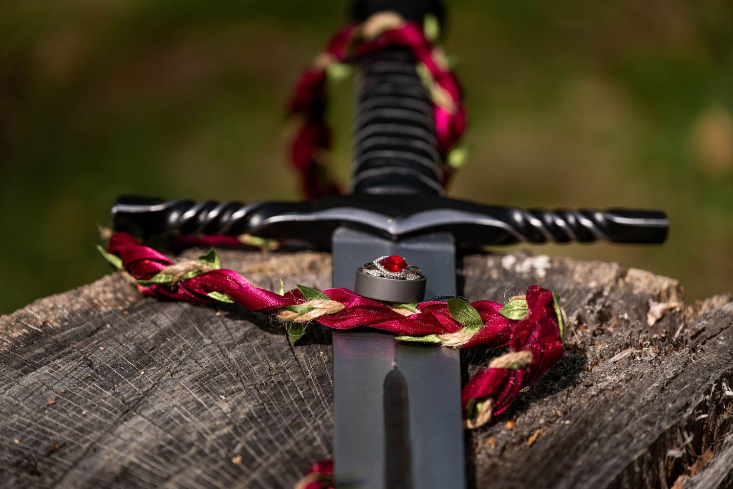 A medieval black sword with a red and gold braided wreath resting on a wooden log outdoors.
