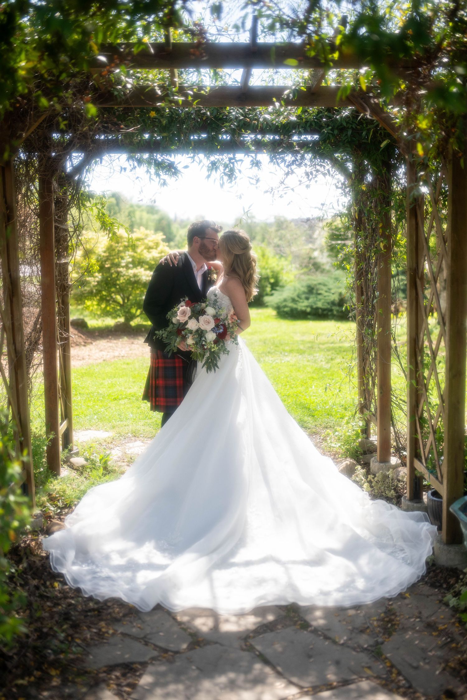 Bride and groom kissing under a vine-covered garden arbor, bride in white gown holding floral bouquet, groom in red tartan kilt.