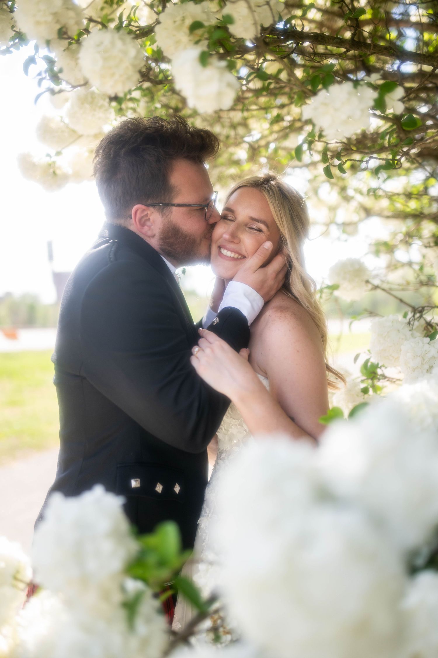 A groom in a kilt kisses his smiling bride on the cheek surrounded by white blooming flowers outdoors.