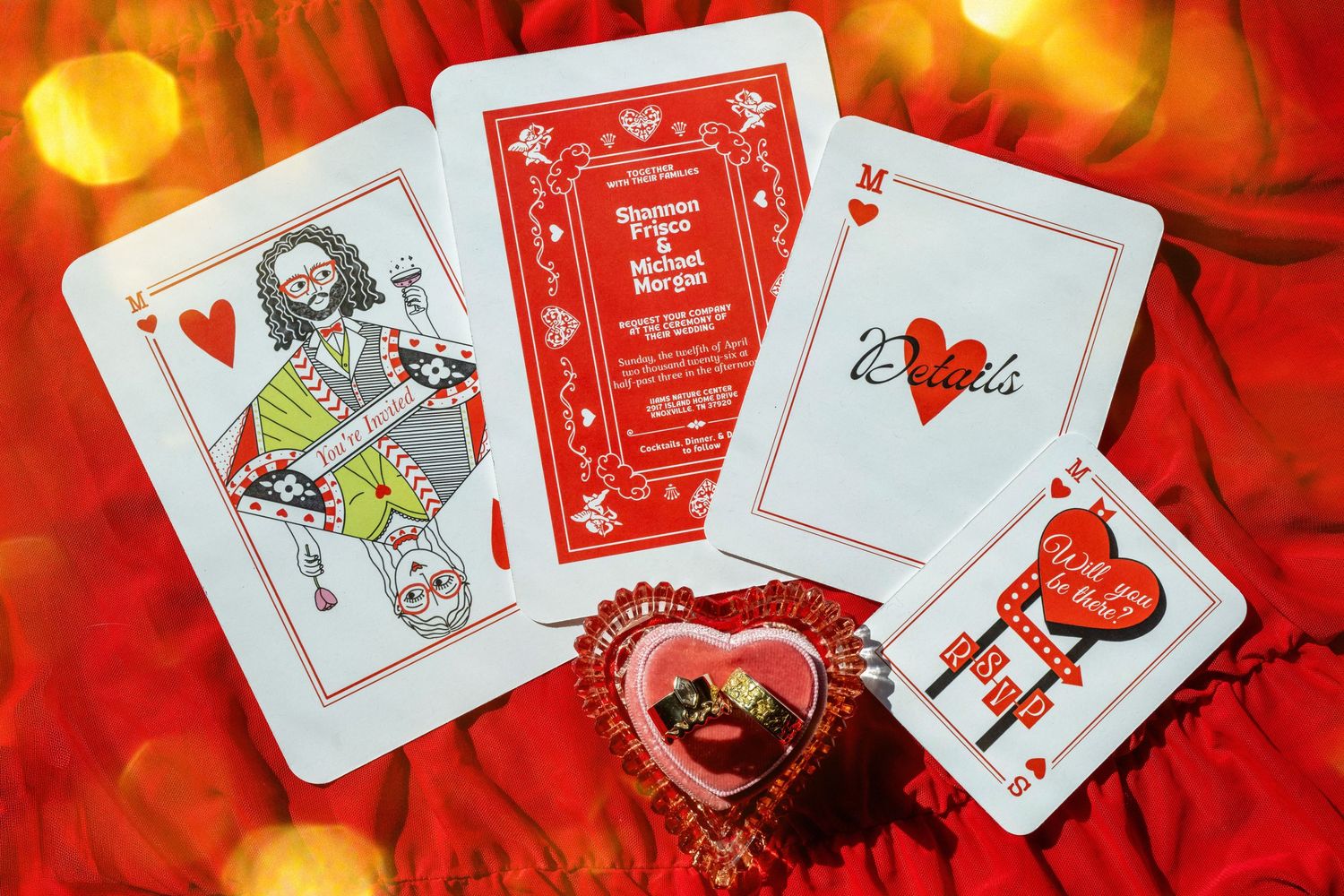 Red playing cards with hearts suit, custom wedding invitation design, and heart-shaped chocolate box on red fabric with bokeh lights.