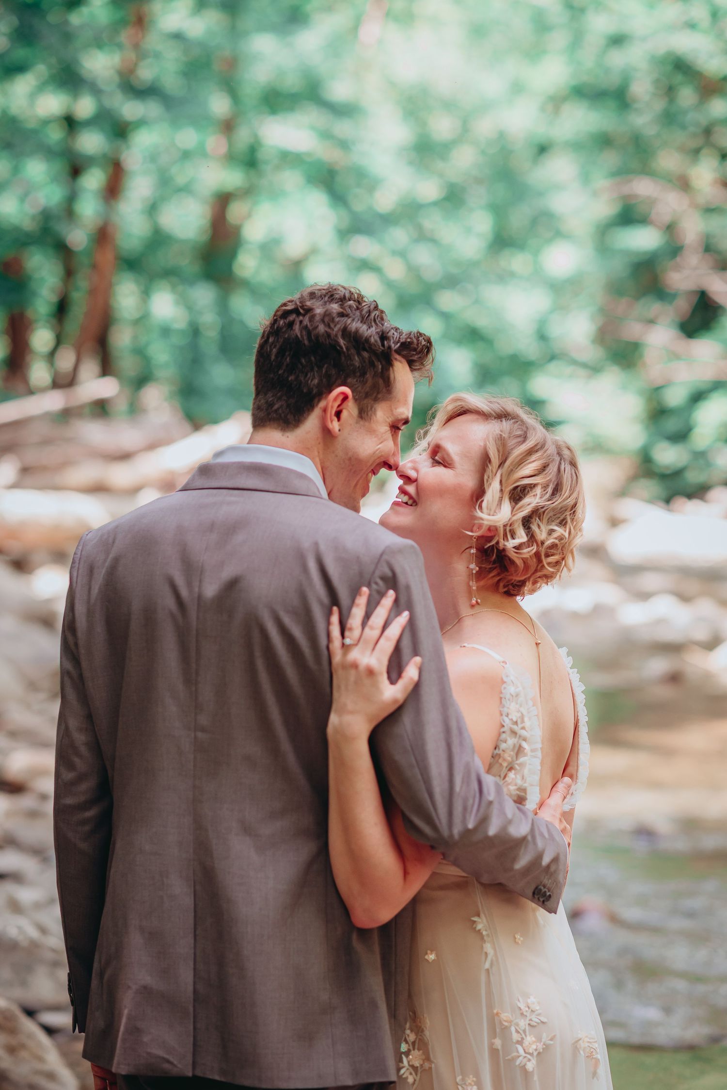 Couple shares intimate moment in ethereal forest setting during wedding photoshoot near stream with soft natural lighting.