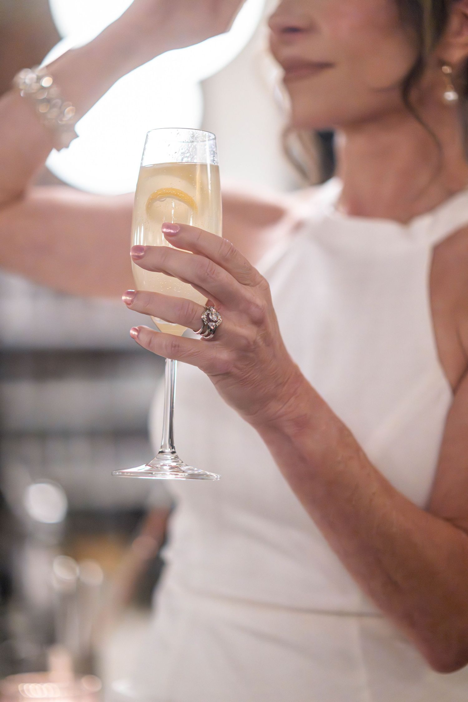 Someone in a white dress holds a champagne flute with a delicate engagement ring sparkling on their hand.
