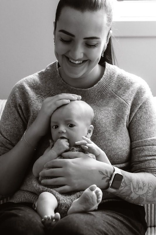 Svart-hvitt bilde av nybakt mor med sin nyfødte gutt sittende på fanget. Mamma holder han på hodet og smiler.