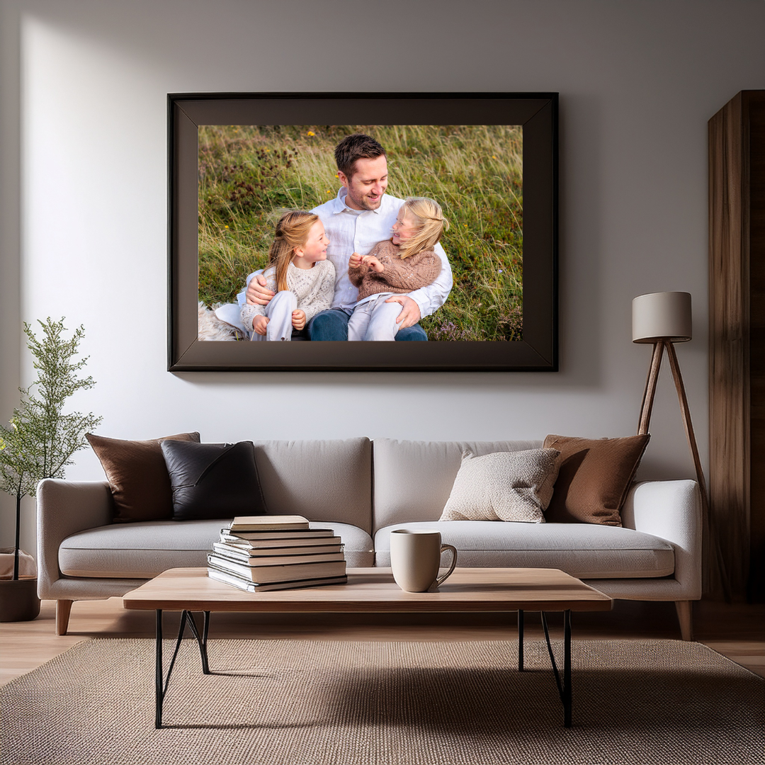 Mockup bilde av et veggbilde av far og to døtre i gresset på Sola over et bord med kaffe og bøker. Reklame for Ørstavik Foto sine produkter.