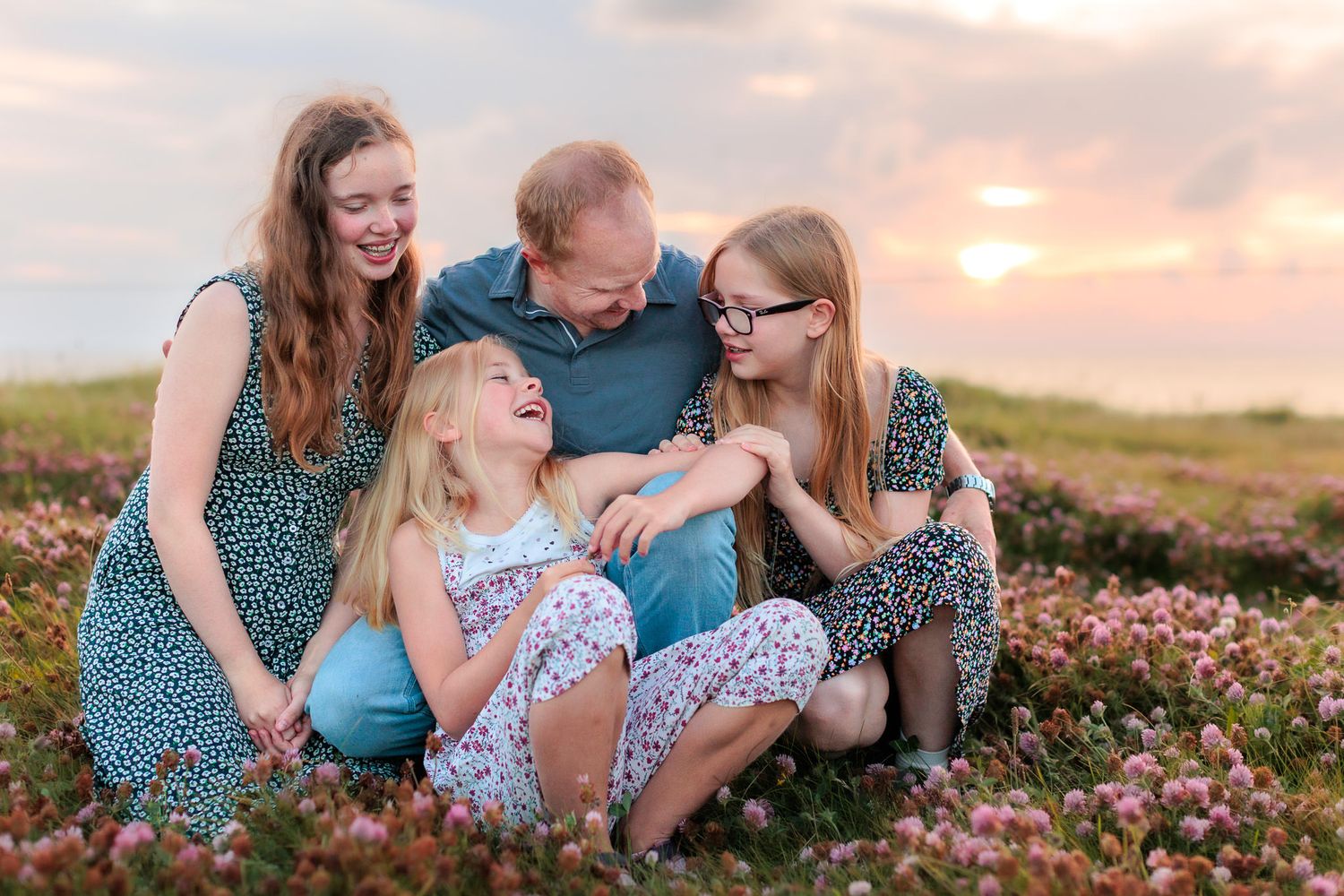 Far og tre døtre sitter tett en eng med nydelige blomster, i solnedgang. Tatt av familiefotograf Ørstavik Foto.