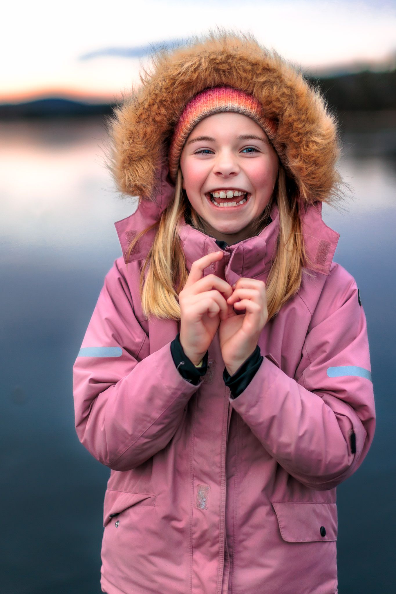 Jente med pelshette smiler til kamera på islagt vann i solnedgang om vinteren. Tatt på Naglestad i Agder.