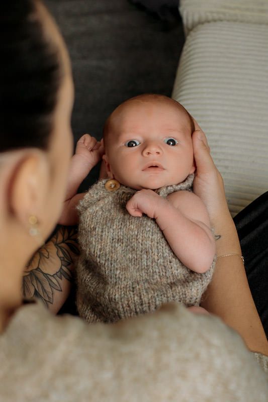 Nyfødt gutt ligger på ryggen i armene til sin mamma og ser på kameraet med store øyne.