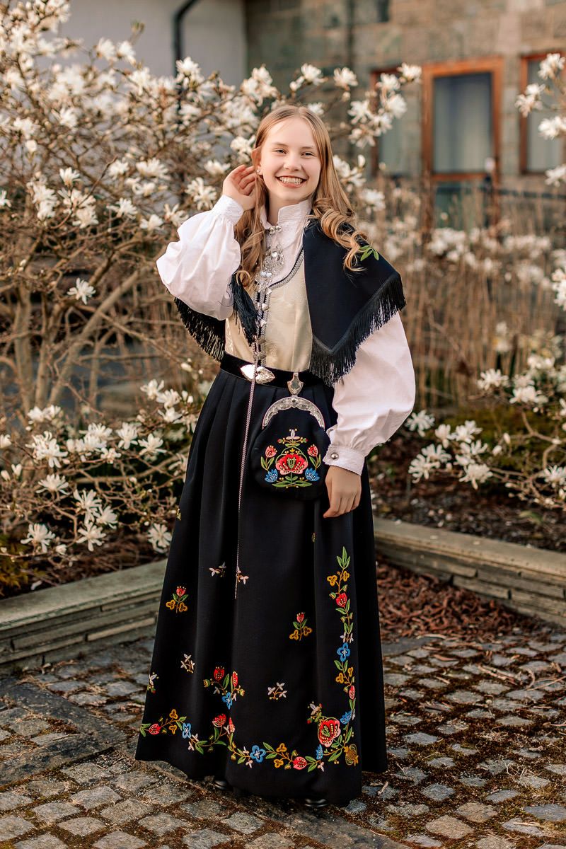 Konfirmant i Jelsabunad med hvit vest står foran et tre med hvite blomster mens hun stryker hånda bak øret og smiler.