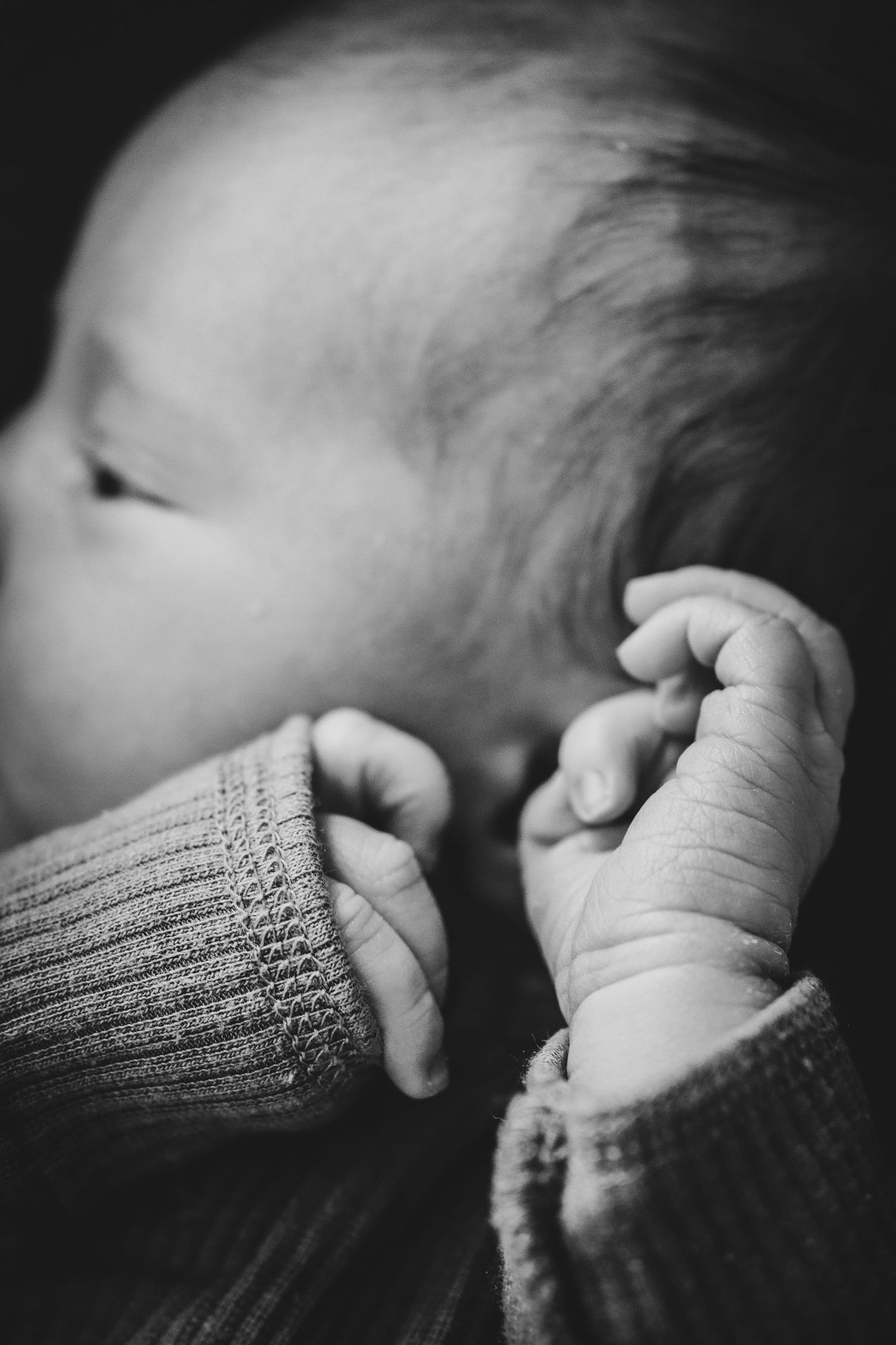 Black and white photo of a sleeping newborn baby with tiny hands raised near their face, wearing a knit sweater.