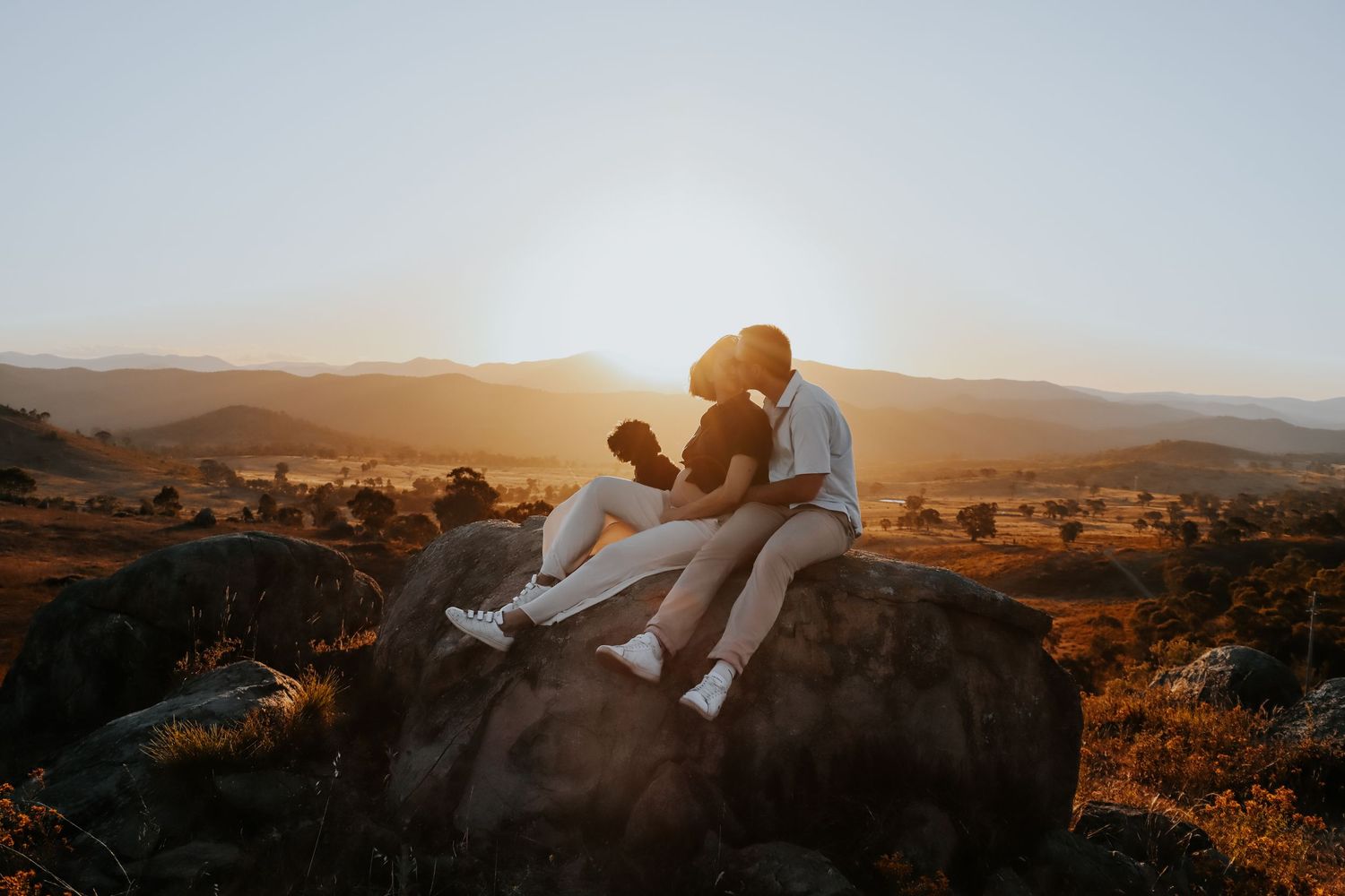 Couple sitting on a rock at sunset with golden light and scenic mountain valley landscape in the background.