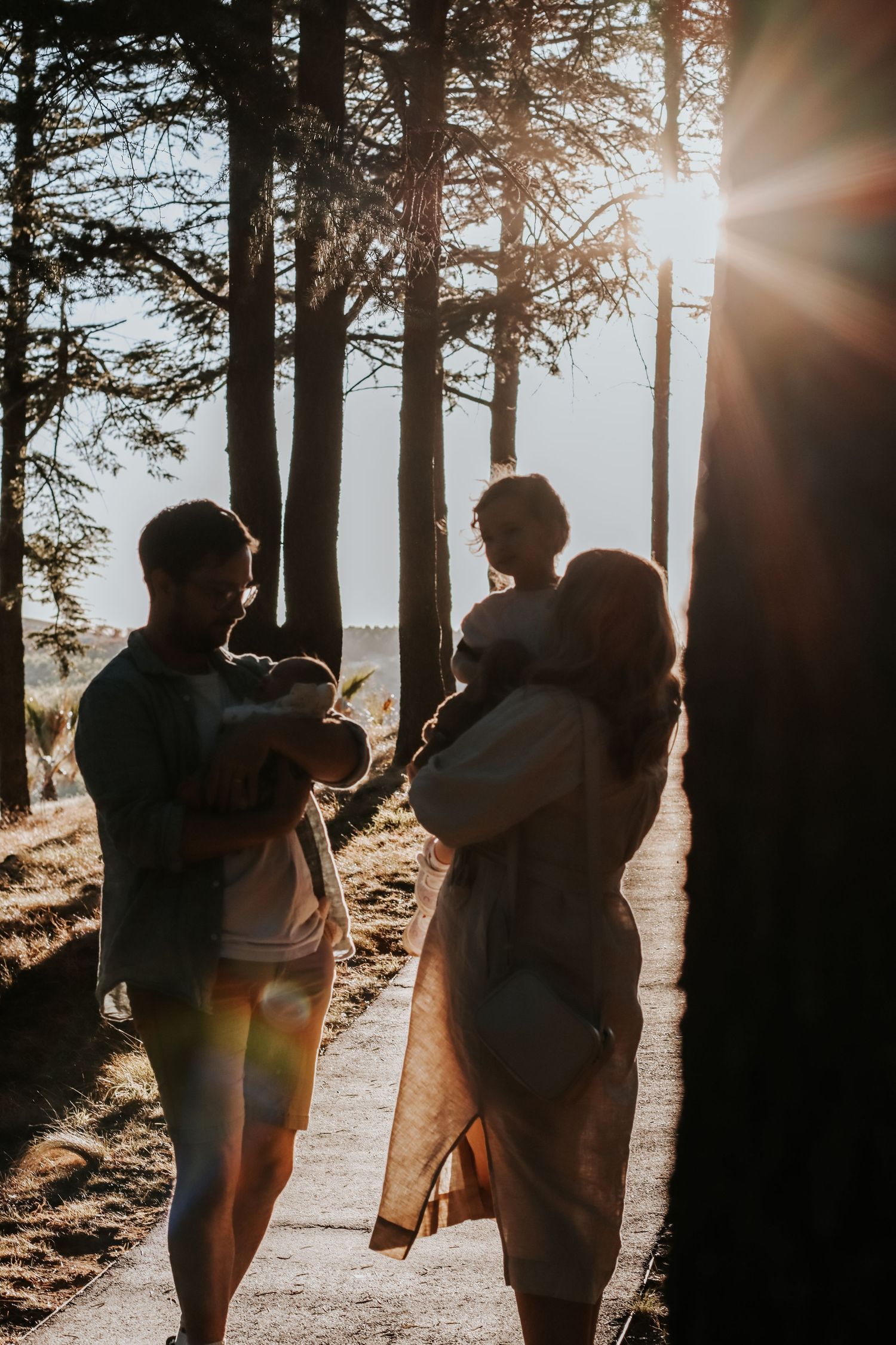 Family silhouetted against golden sunset light in forest, parents holding children on woodland path.