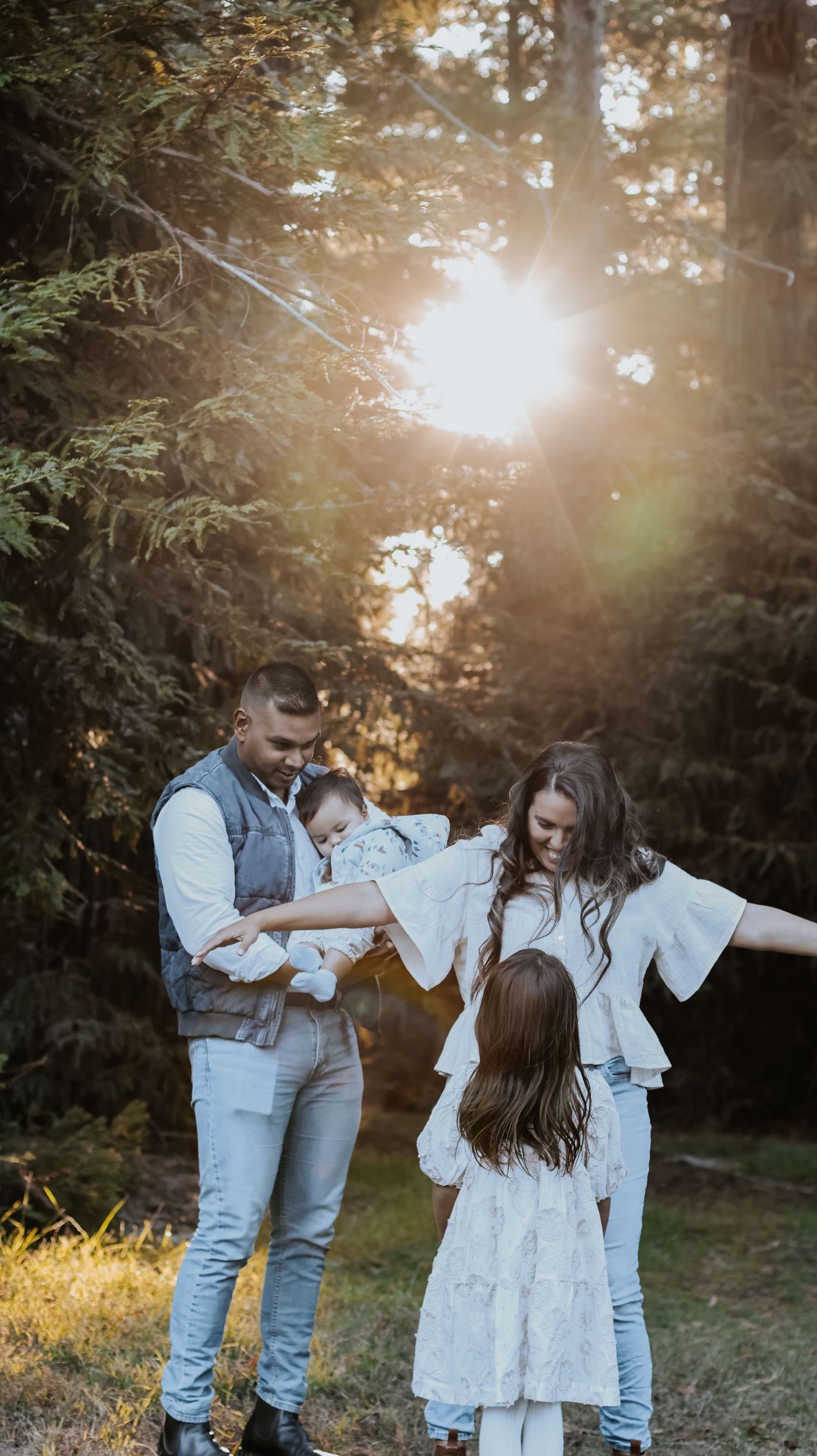 Family of four enjoying a golden sunset in a lush forest, parents playing with young children in white outfits.